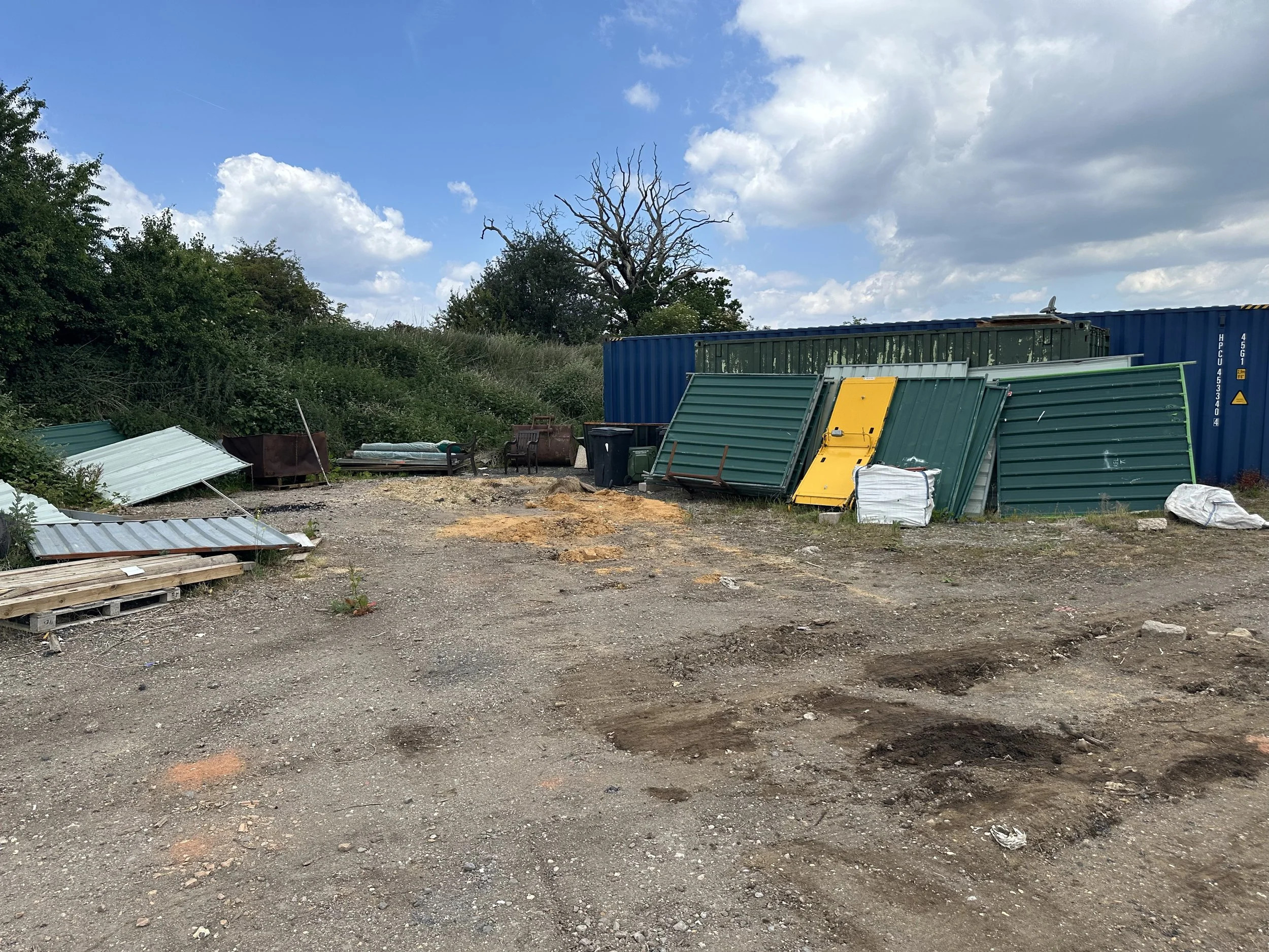 A messy outdoor area with a dirt ground, scattered metal sheets, a blue shipping container, various discarded items, and a leafless tree against a cloudy sky.