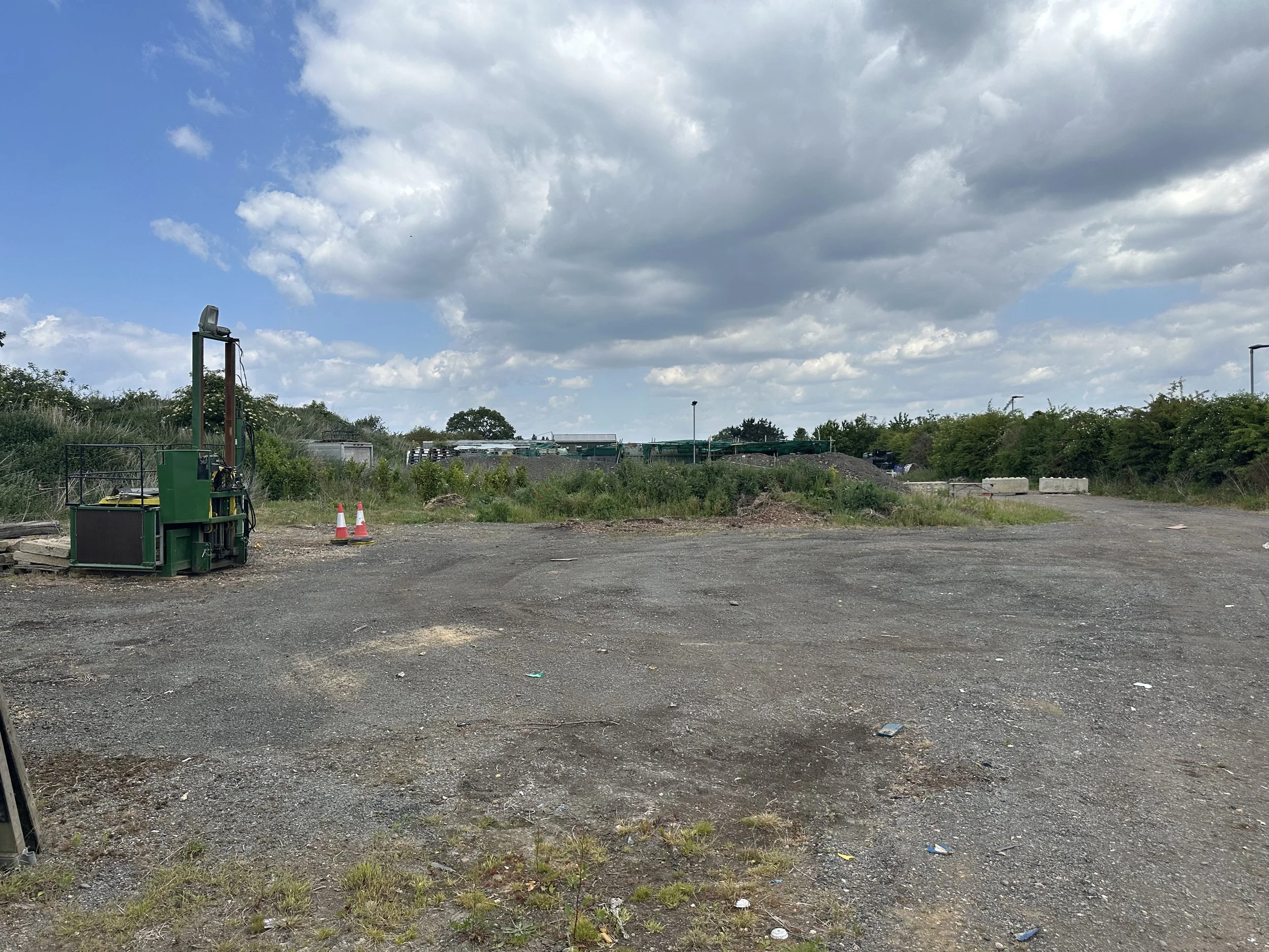 Open lot with gravel and dirt, some construction equipment, orange traffic cones, grassy patches, bushes, trees, and a cloudy sky.
