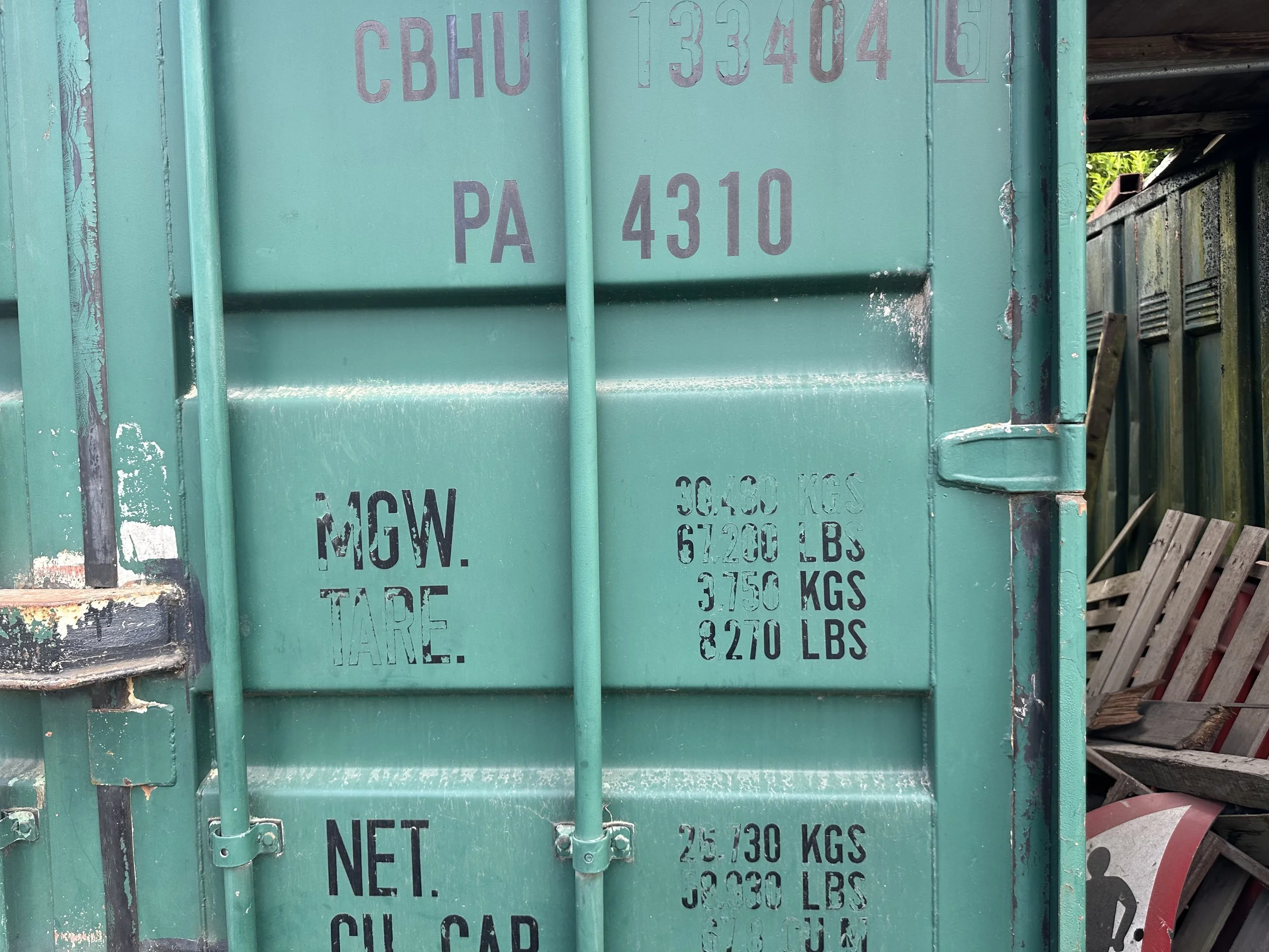 Close-up of a green metal container with faded black stenciled text showing weight and specifications, covered by vertical bars.
