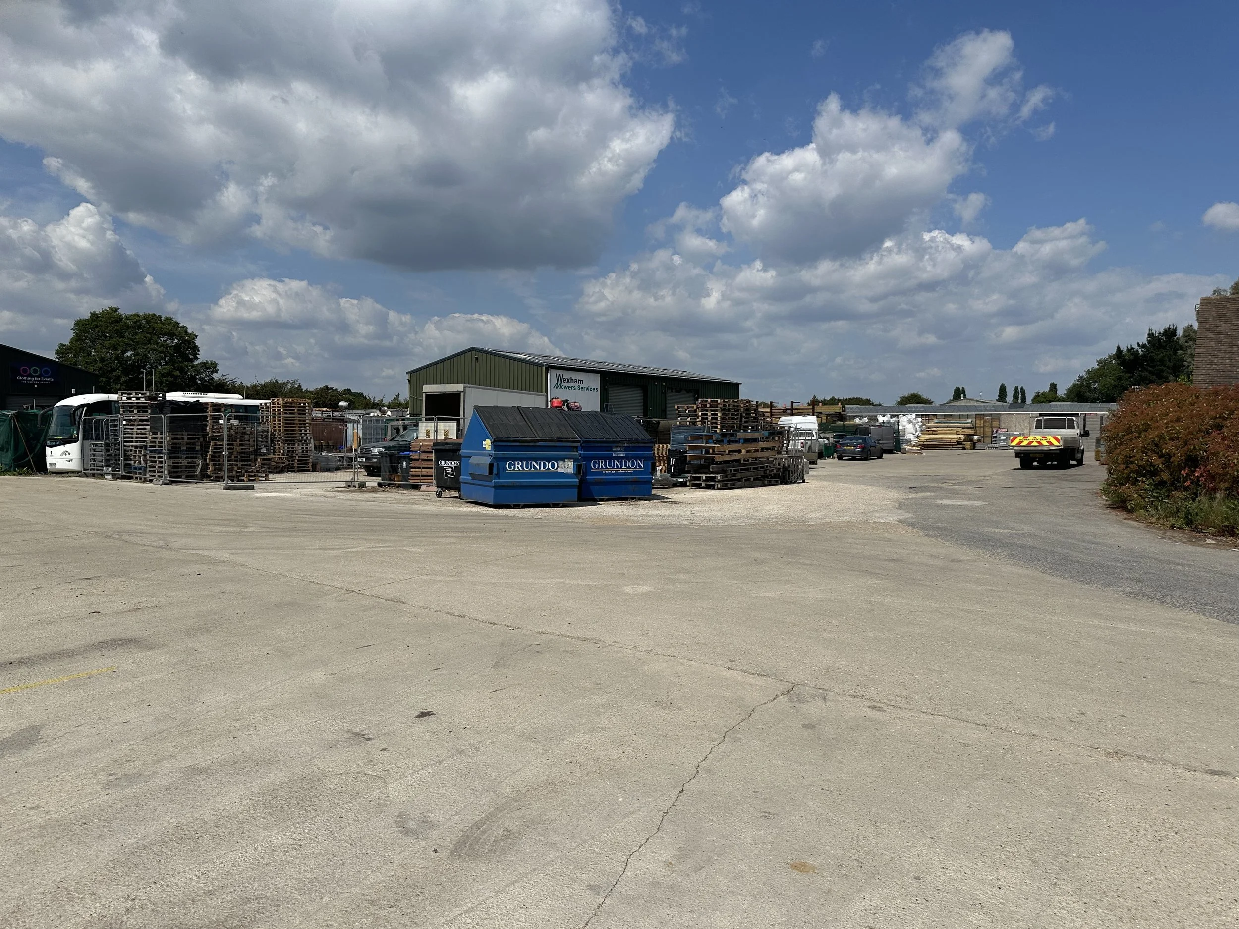 A yard or storage area with various pallets, metal containers, and vehicles, including a bus and a white truck, under a partly cloudy sky.