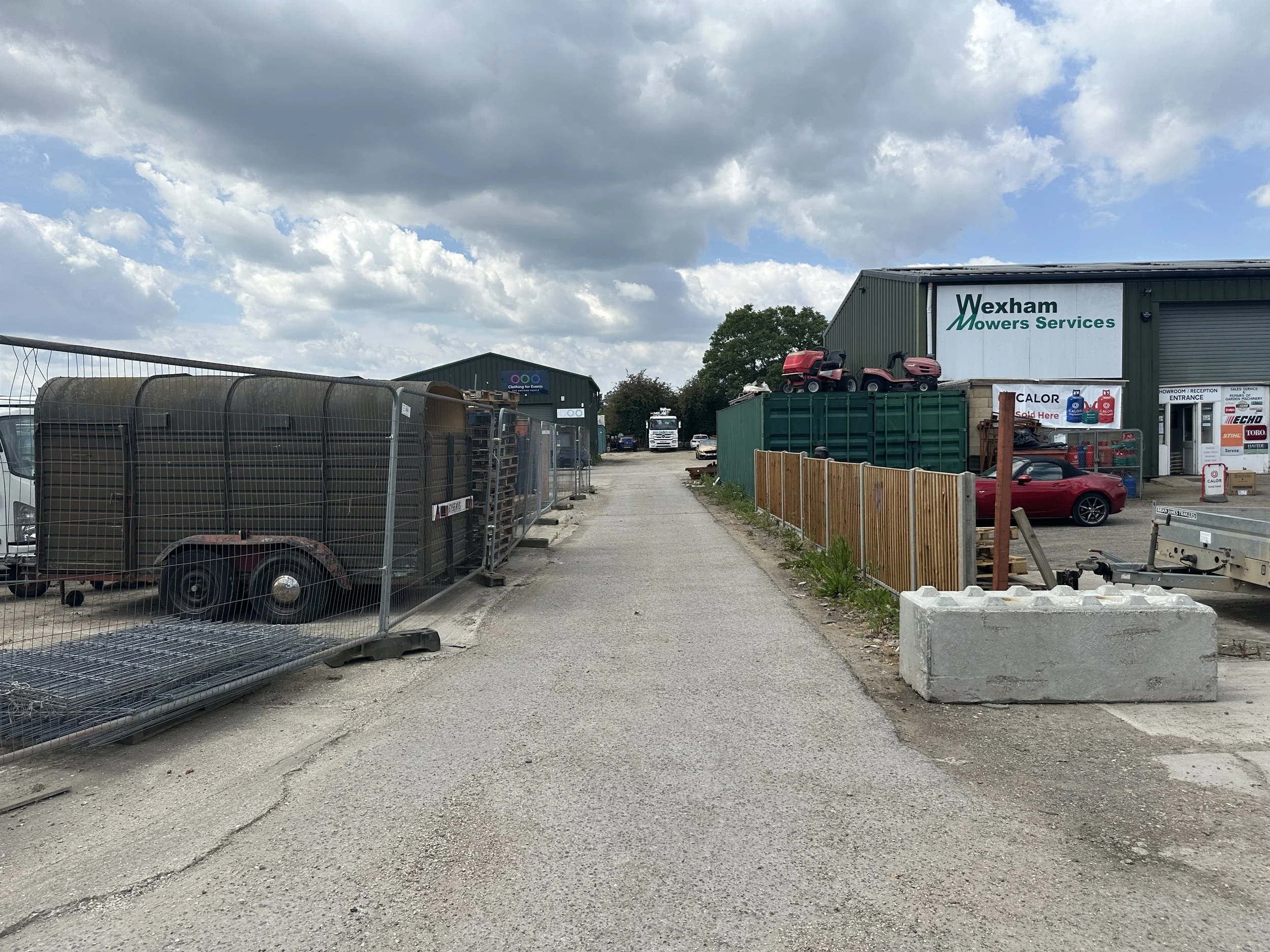 A storage yard with metal fencing, a gravel pathway, and various equipment and vehicles, including lawnmowers on a trailer and a red sports car, outside a building labeled 'Wexham Mowers Services' under a partly cloudy sky.