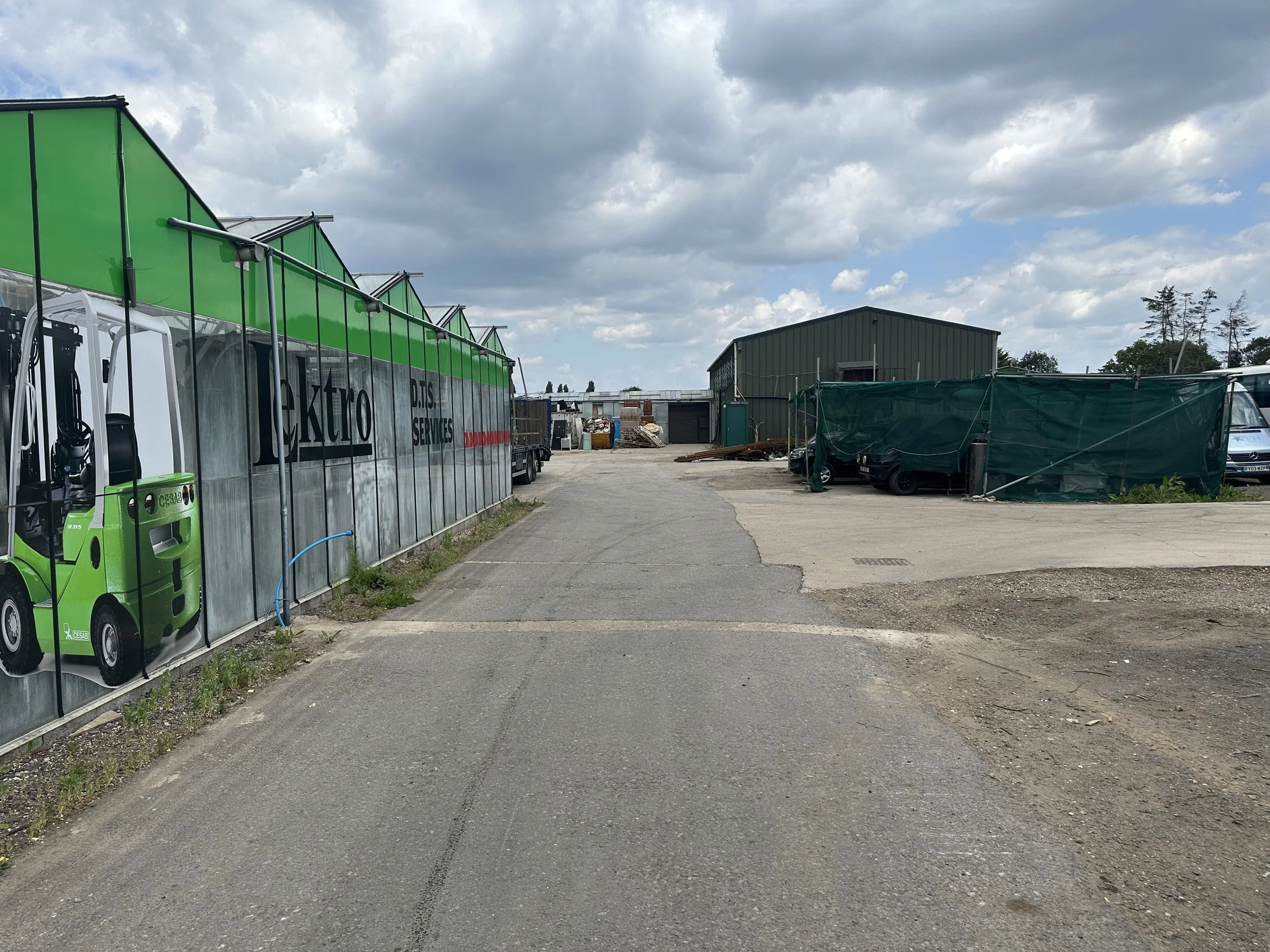 An industrial area with a large green and gray vehicle on the left side, a green warehouse, and a storage area with materials and equipment. The sky is partly cloudy.