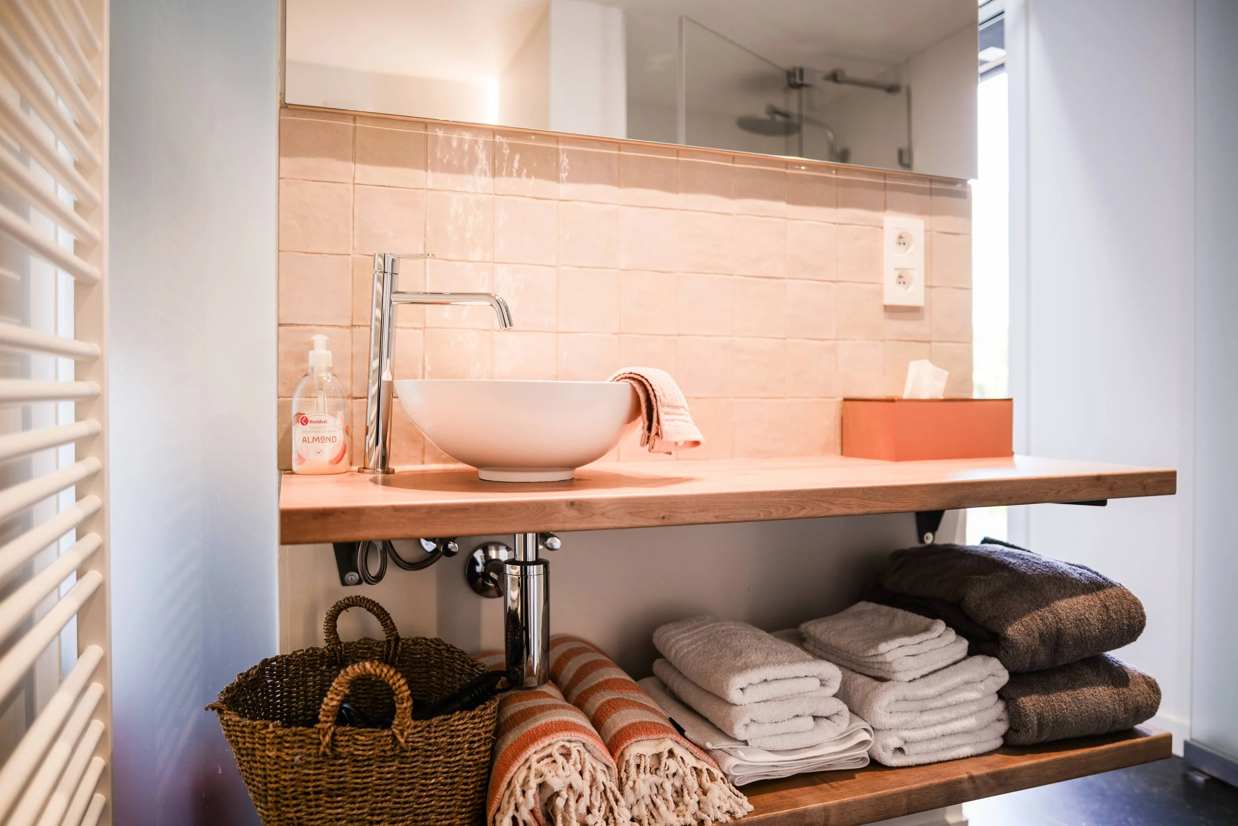 A bathroom vanity with a round white sink and a modern chrome faucet. There is a bottle of hand soap beside the sink. Below the vanity, there are neatly folded towels, rolled-up blankets, woven baskets, and a dark brown pillow. The background feature