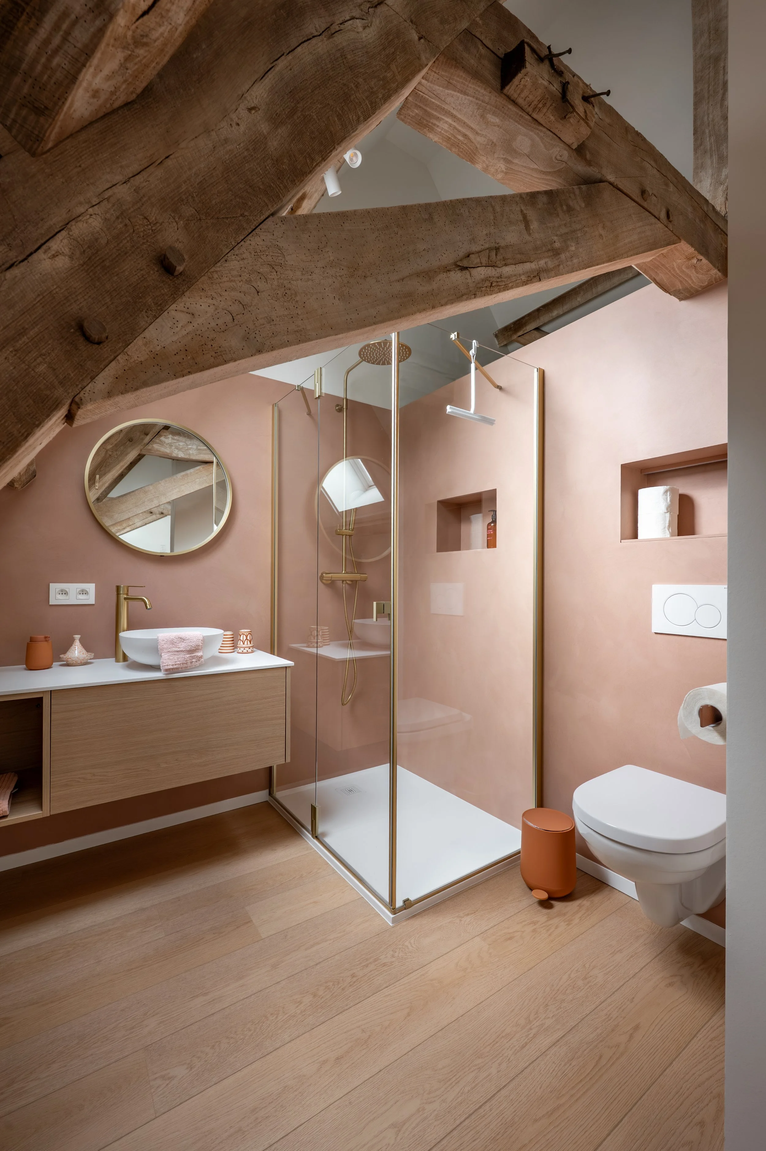 A modern bathroom with pink walls, wooden floor, a round mirror, a white sink with a gold faucet, a glass shower enclosure with gold fittings, a wall-mounted toilet, and wooden beams on the ceiling.