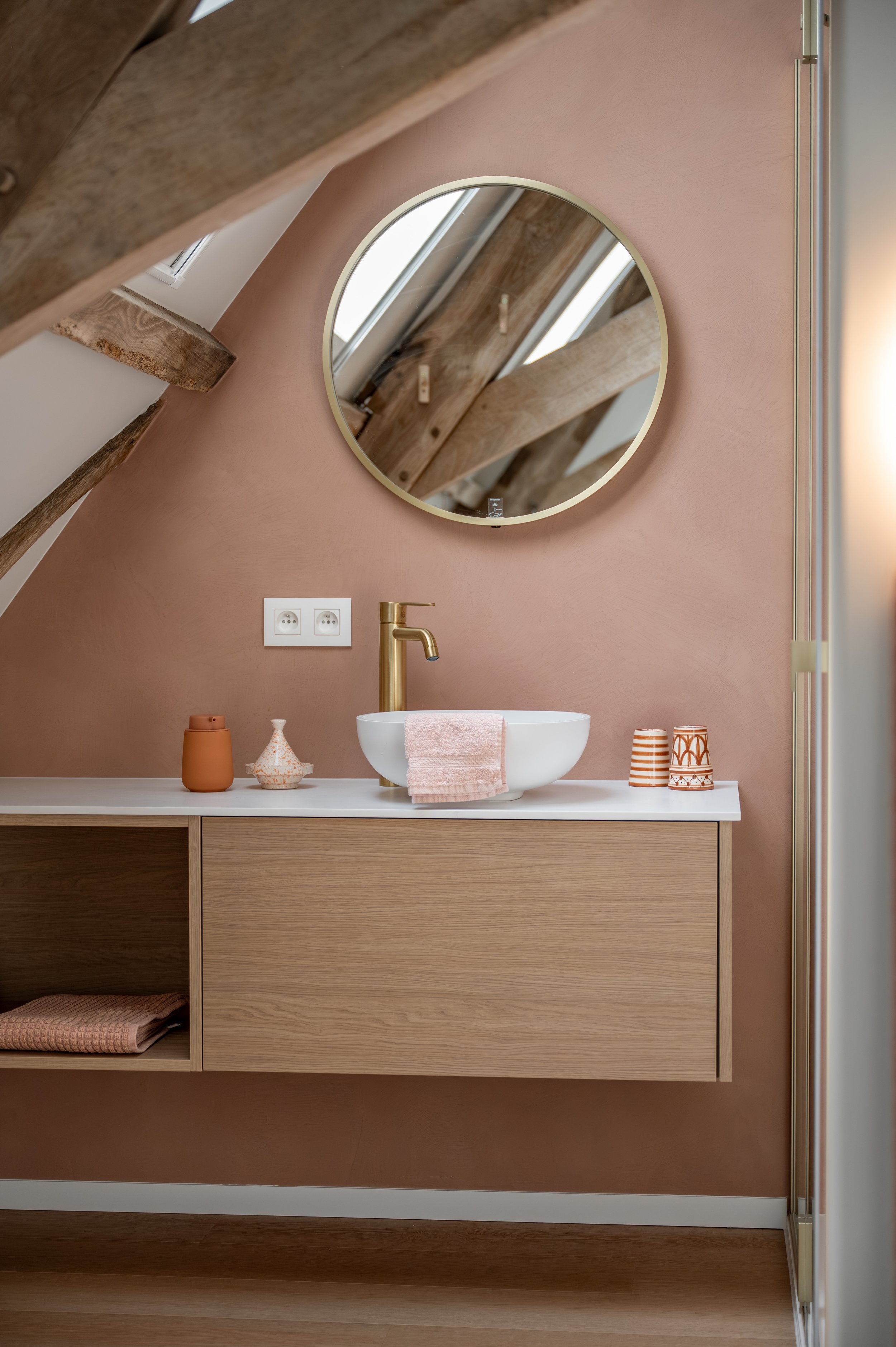 A modern bathroom vanity with a round mirror, wooden accents, a white vessel sink, a gold faucet, and decorative items on a peach-colored wall.