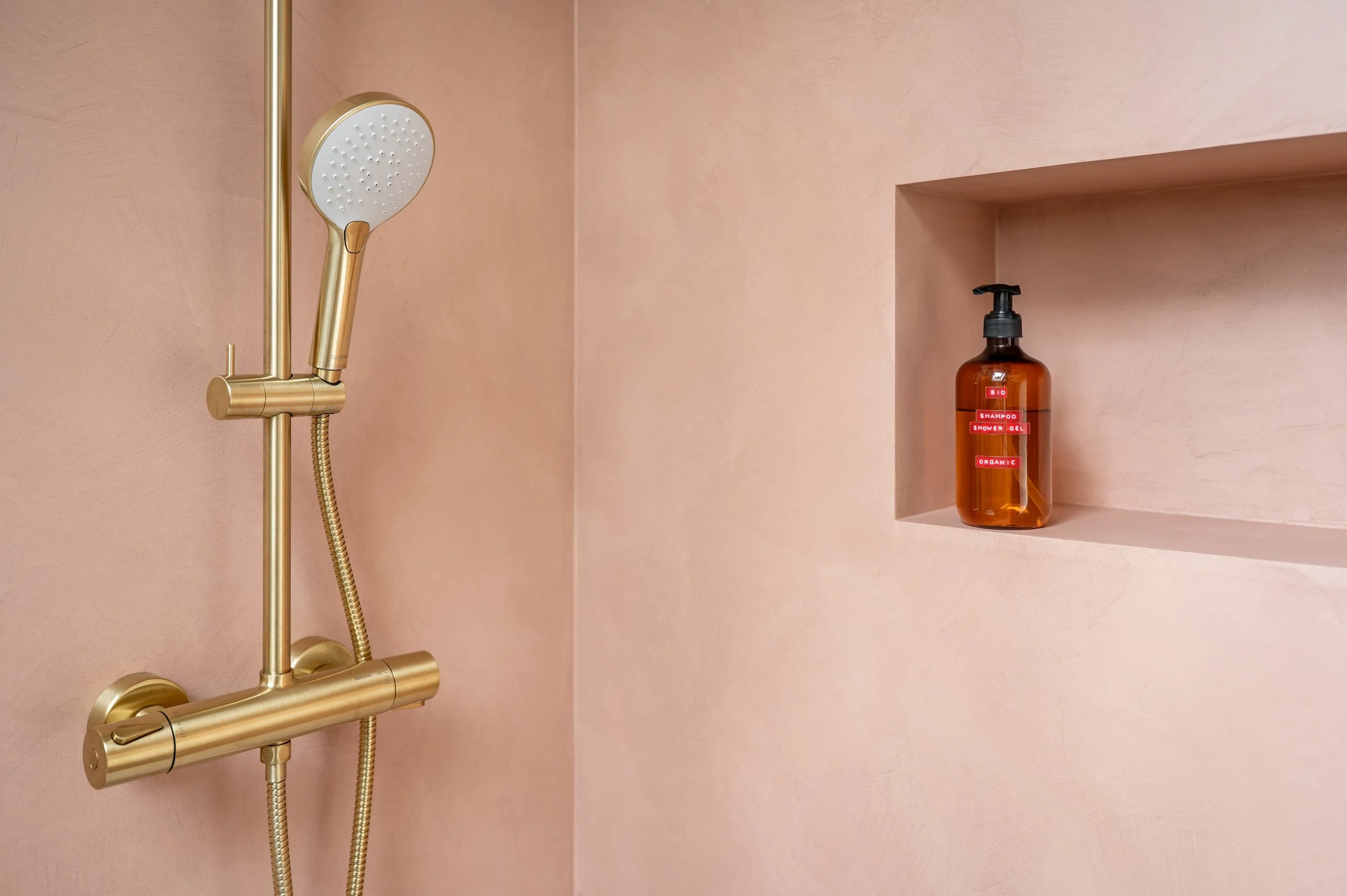 A shower with a gold showerhead and matching faucet, pink walls, and a built-in shelf holding a brown bottle labeled 'Bio Shampoo Shower Gel Organic'.