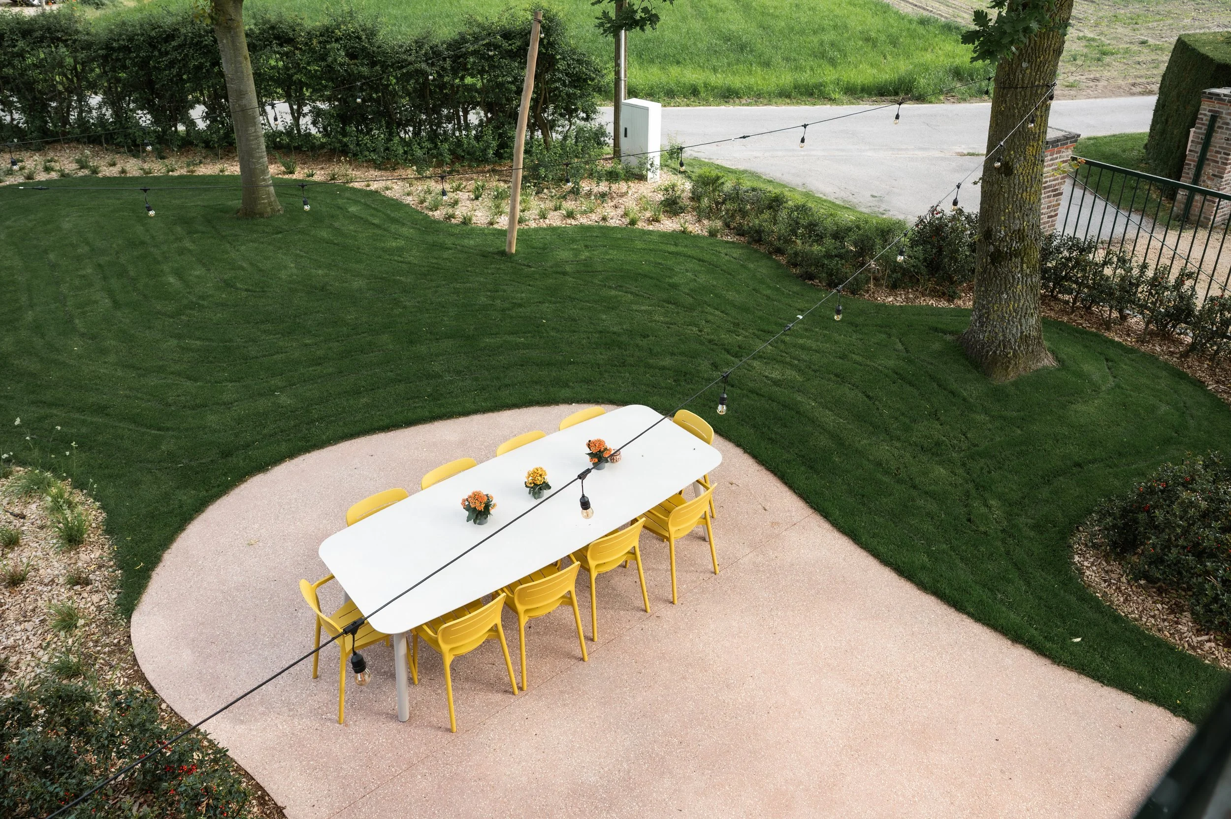 A backyard patio with a white table and yellow chairs, decorated with small flower arrangements, surrounded by a well-manicured lawn and string lights, trees, bushes, and a driveway in the background.