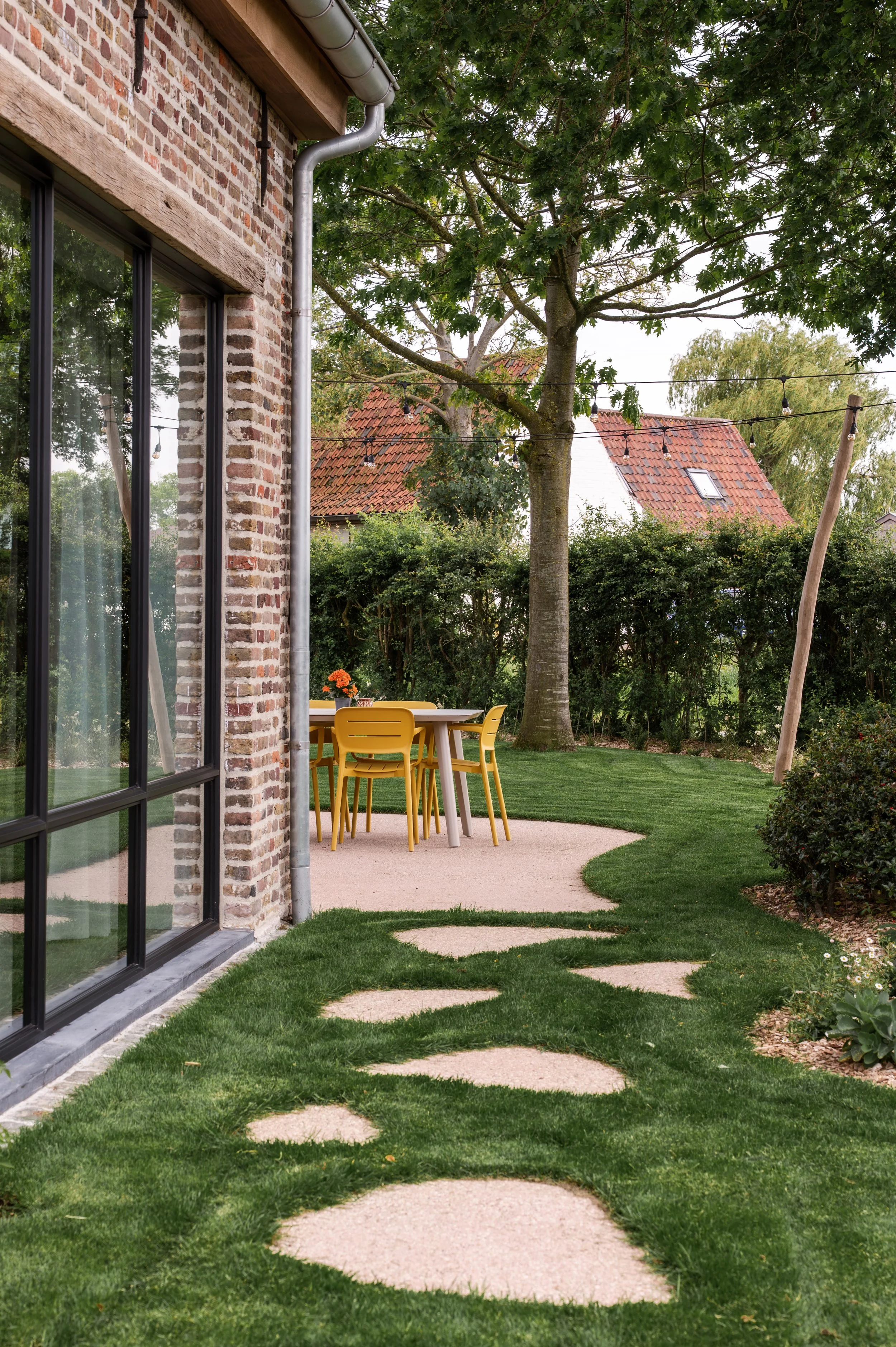A backyard with a brick house wall on the left, a large tree, a yellow outdoor dining set, and string lights. There is a winding concrete path through the green lawn.