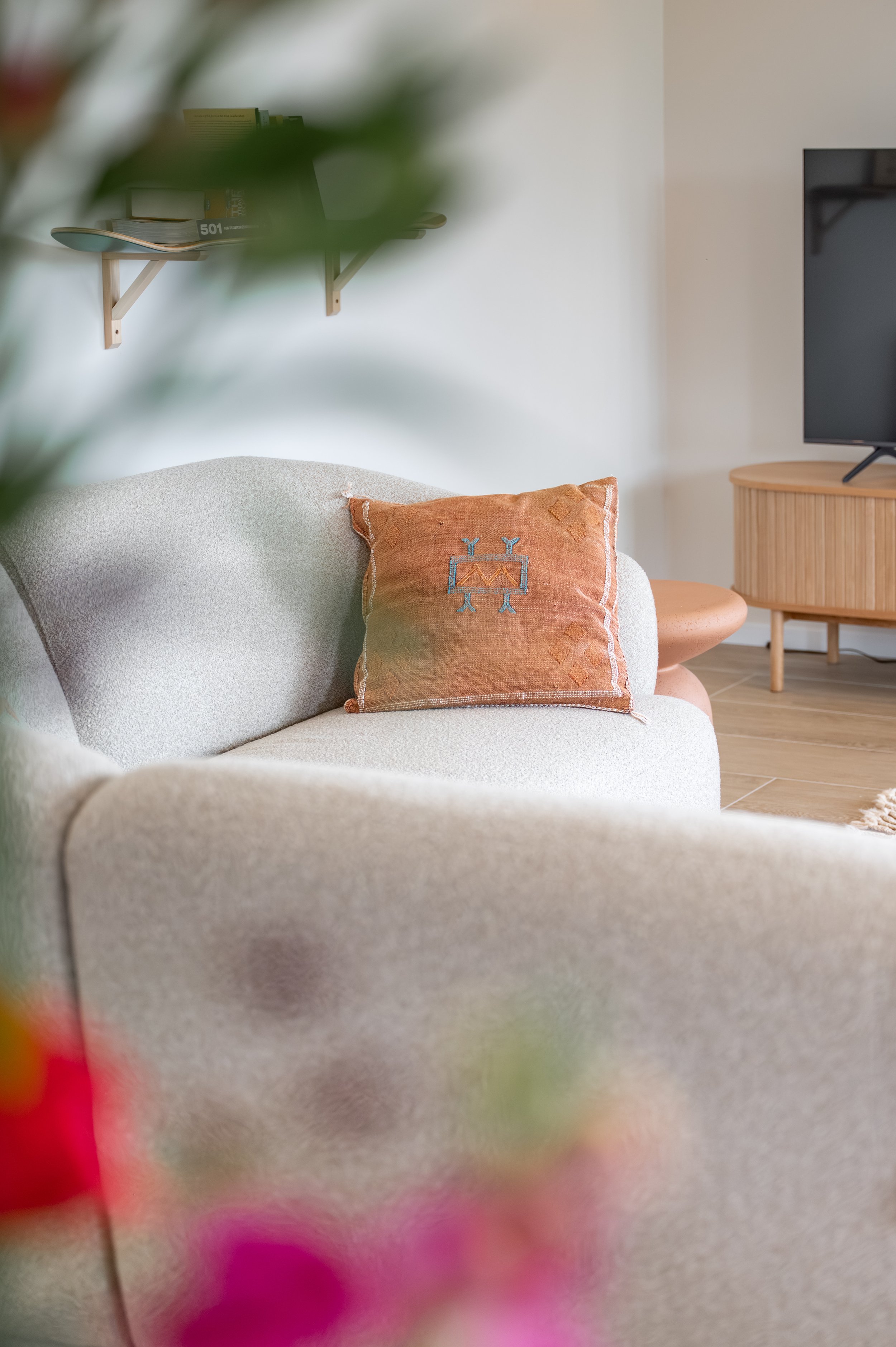 Living room with a beige sofa, a decorative pillow, a TV on a wooden stand, and a floating shelf with books in the background.