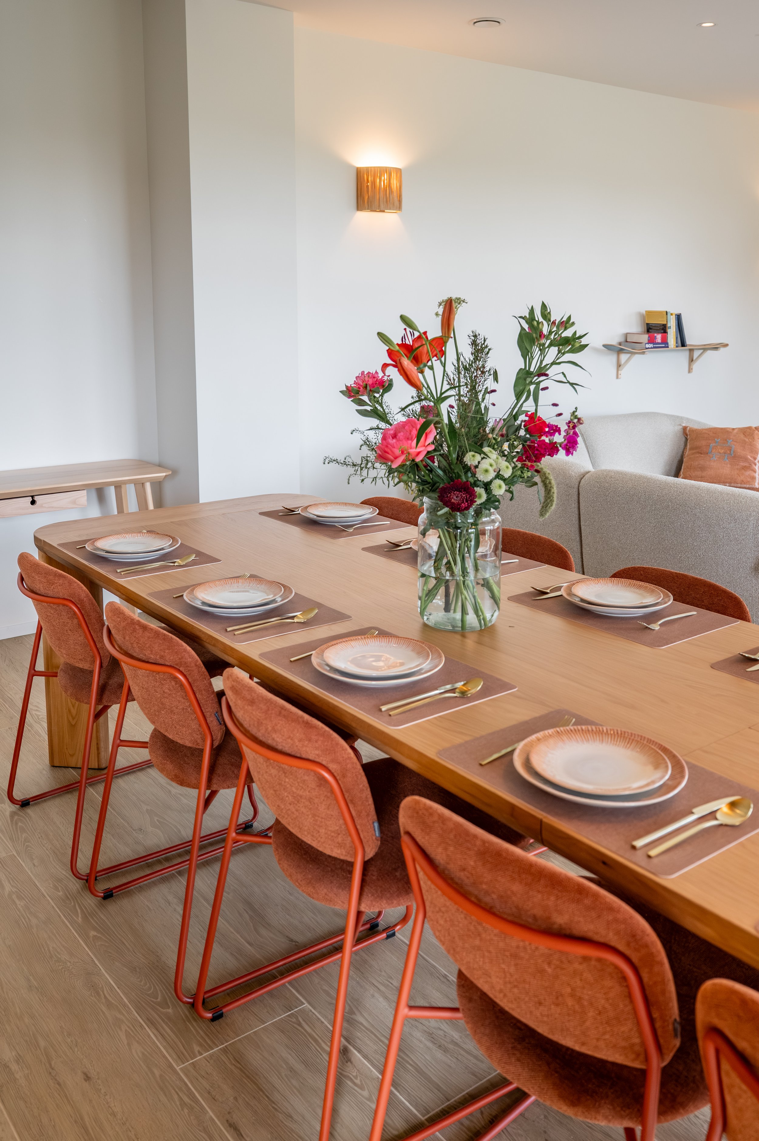 A dining room with a wooden table set with plates, gold utensils, and brown placemats. There are nine burnt orange upholstered chairs around the table. A clear vase with a large floral arrangement sits in the middle of the table. In the background, a