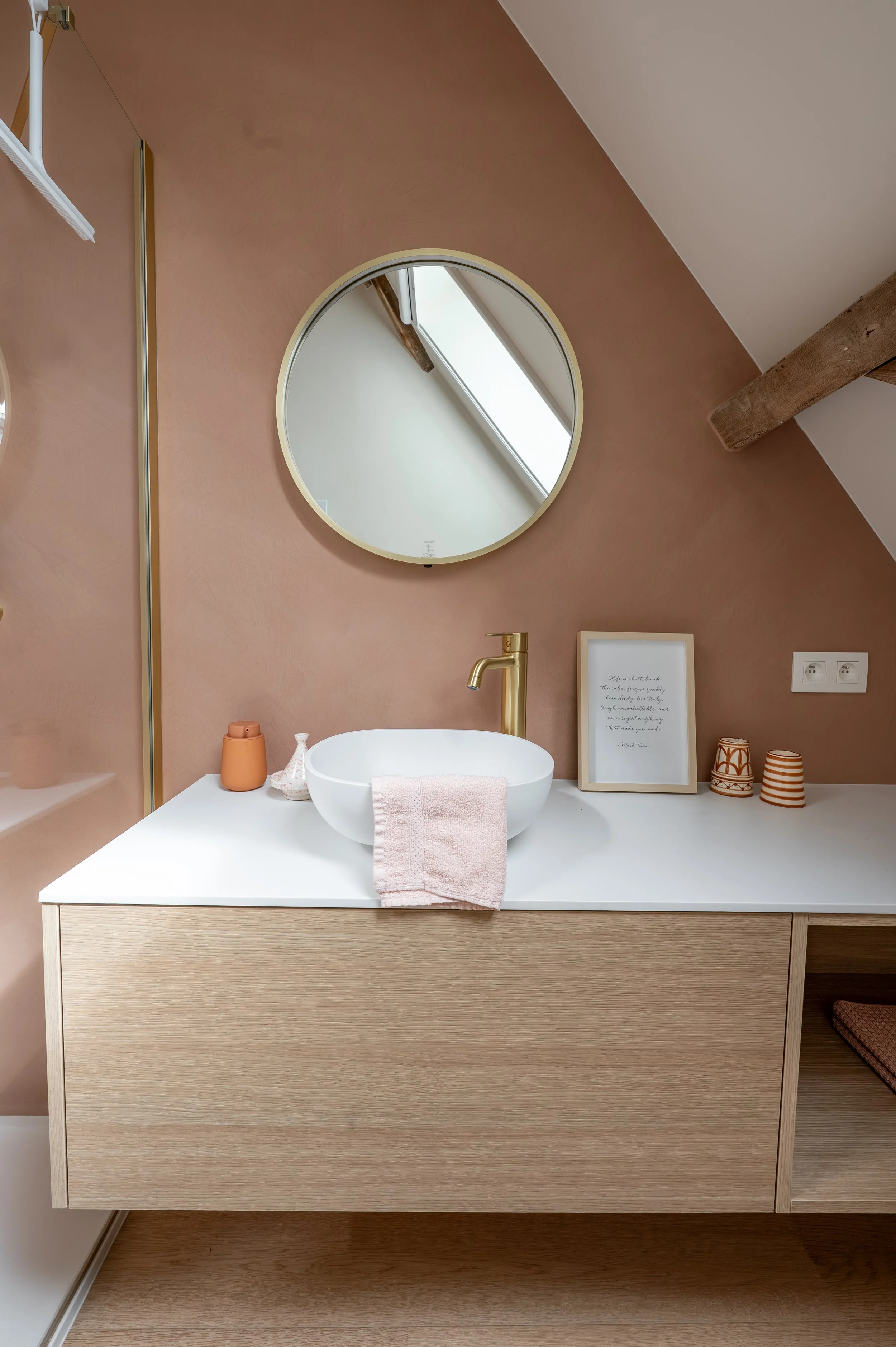 A modern bathroom sink area with a round mirror, a white countertop with a vessel sink, a gold faucet, a pink towel, and decorative items, set against a warm-toned wall with wooden accents.