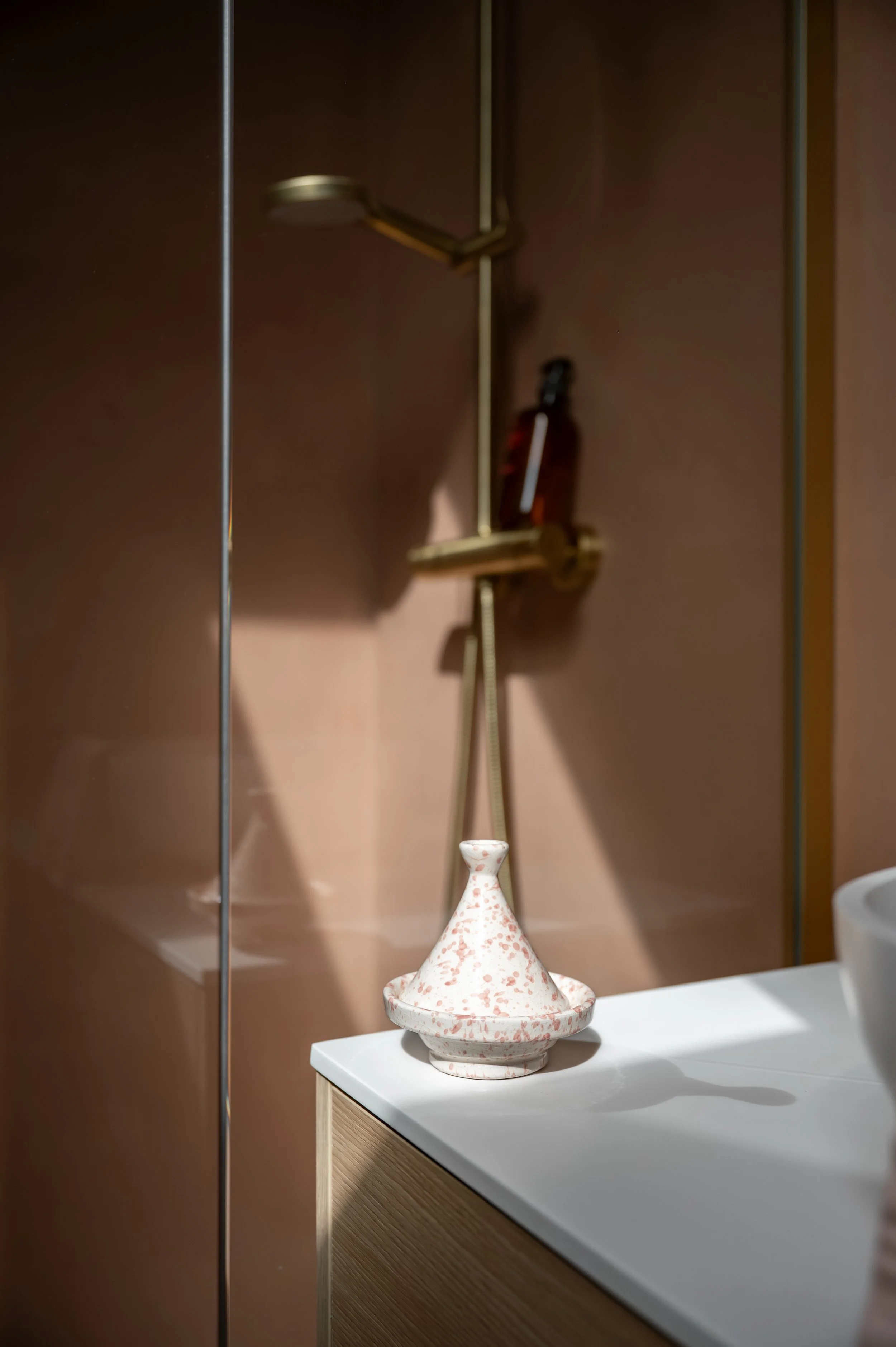 Ceramic vase with a wide base and pointed top, white with pink speckles, placed on a counter with a wood grain front, inside a room with brown walls and a metal shower head in the background.