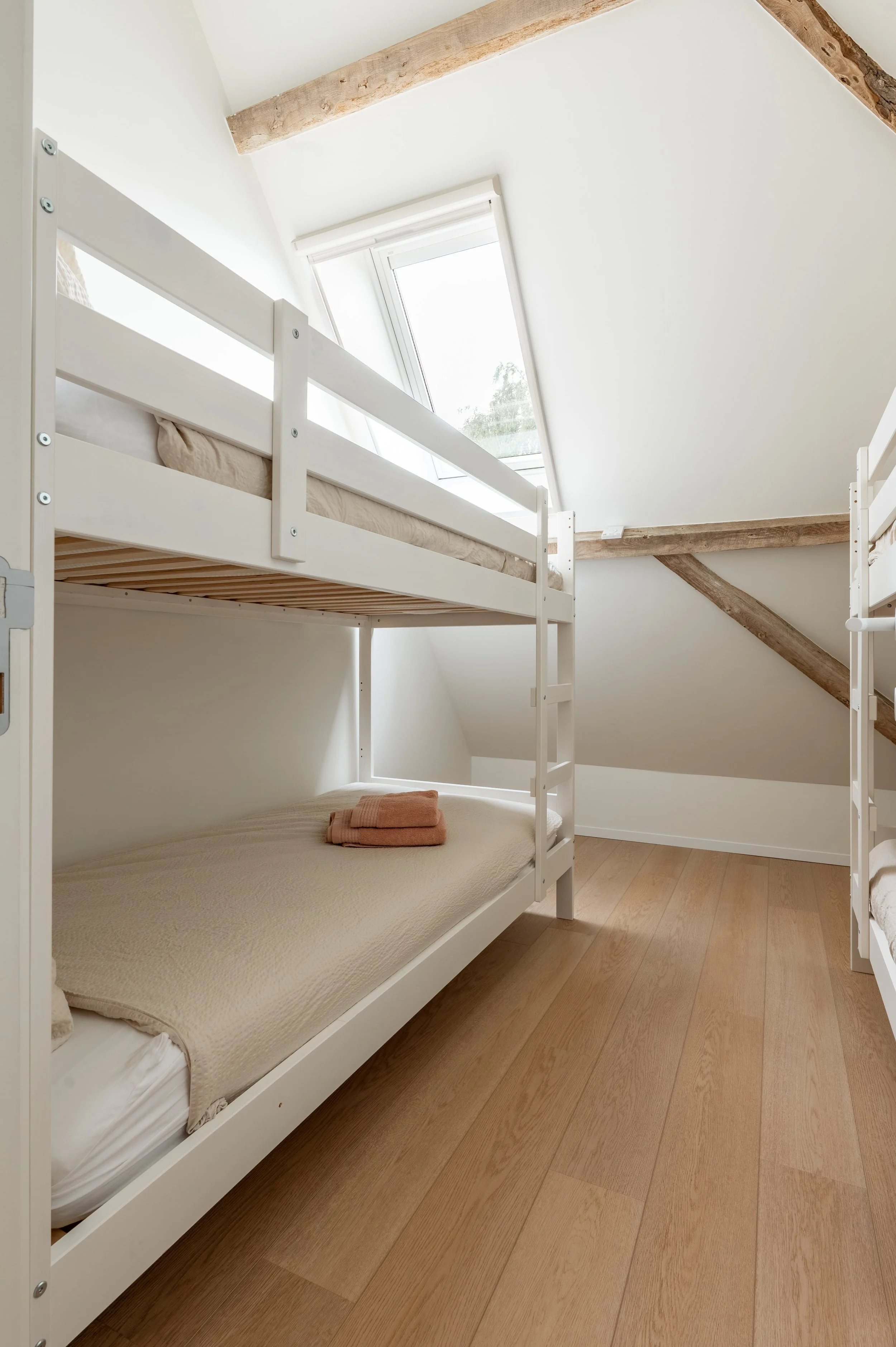 A bedroom with a white bunk bed under a sloped ceiling, featuring a window with natural light and wooden floor.