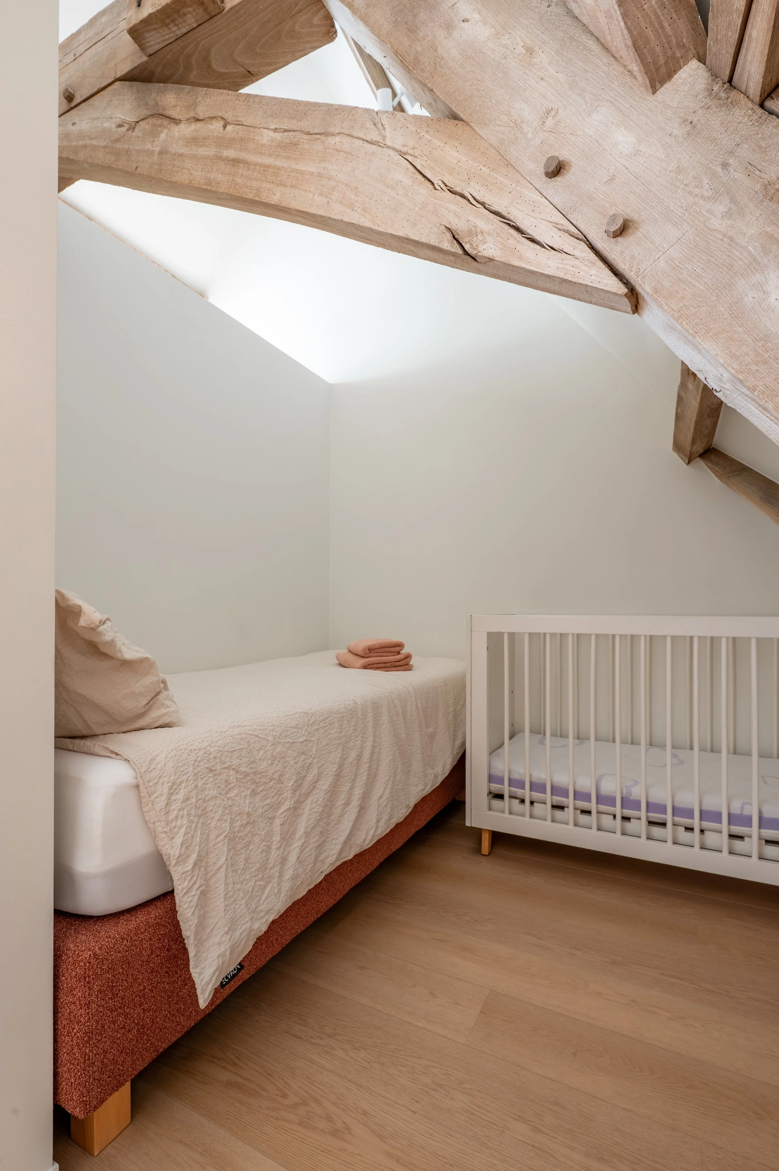 A bedroom with a large wooden beam ceiling, a bed with a beige quilt, a stack of folded towels, and a white crib next to the bed. The flooring is light wood.