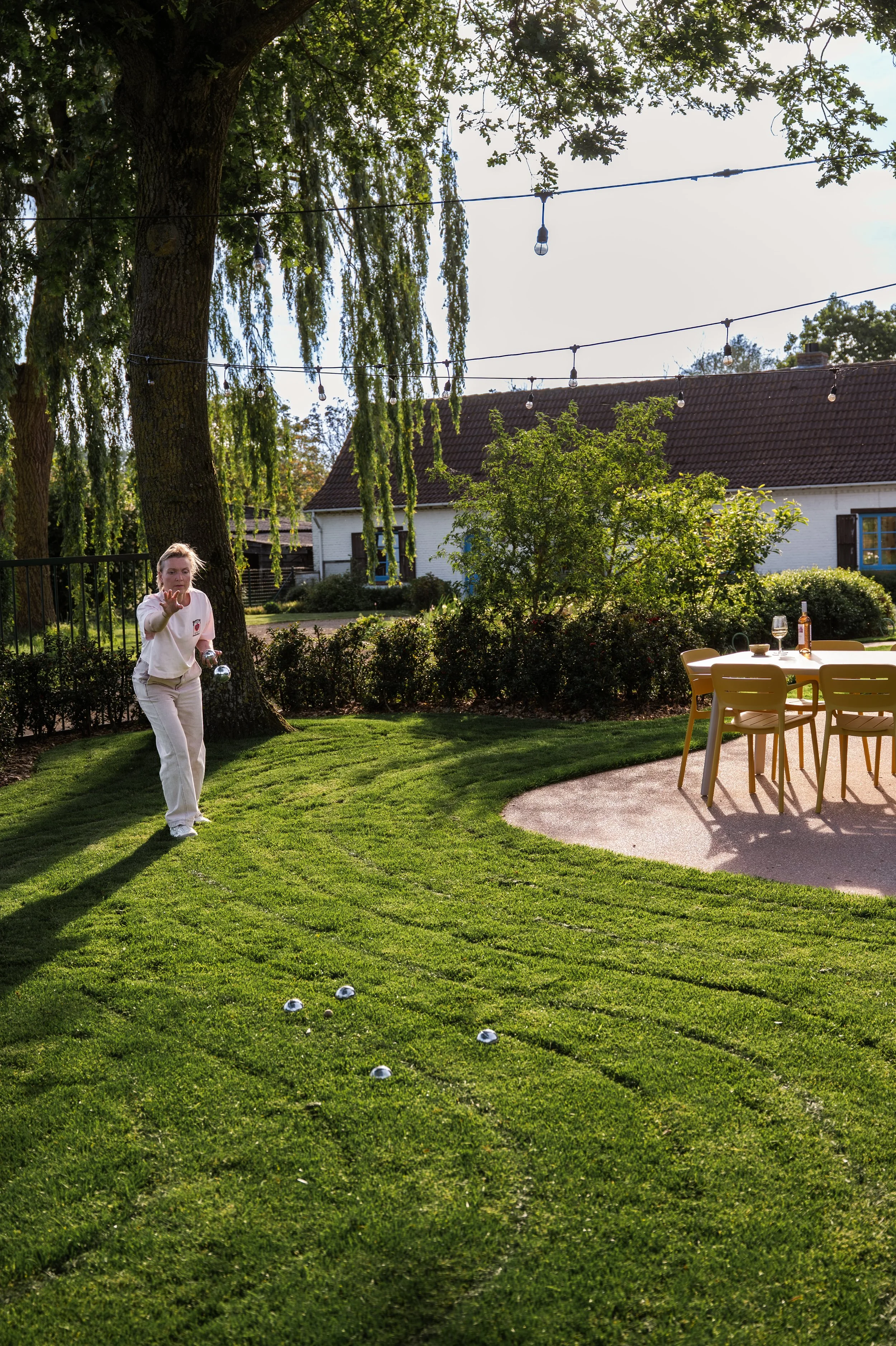 A woman casting a bowling ball in a backyard garden during daytime, with a table set for drinks and a house in the background.
