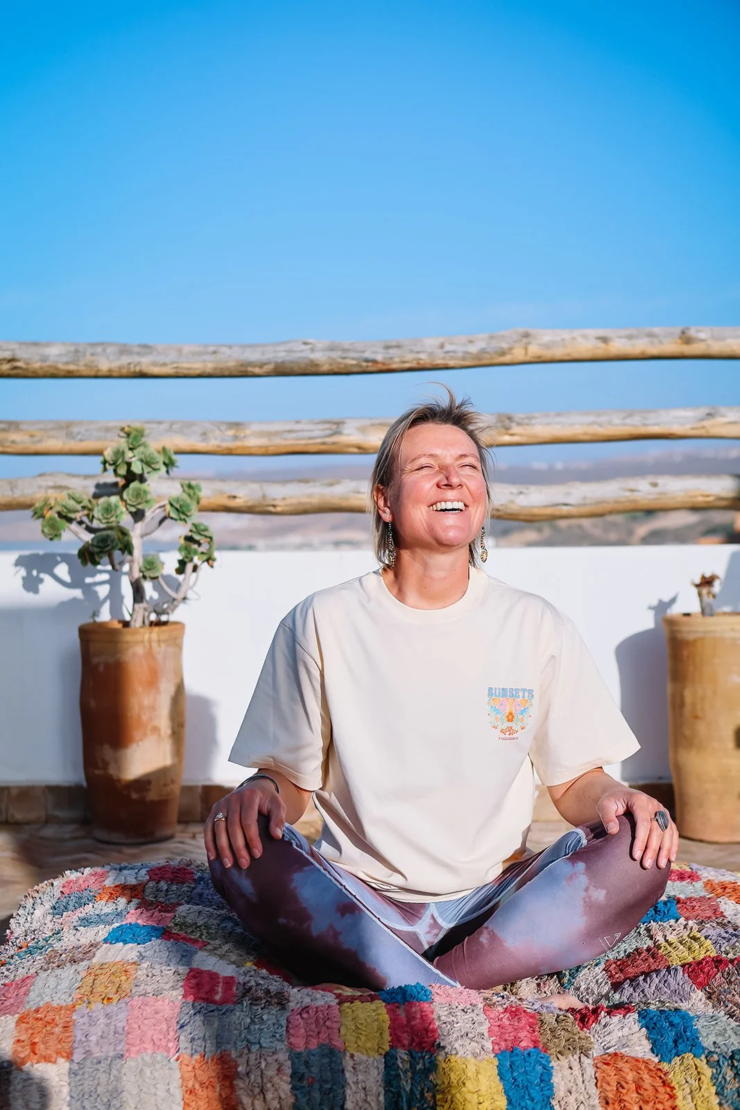 A woman sitting on a colorful blanket outdoors, smiling with her eyes closed, with potted plants and a wooden fence in the background under a bright blue sky.