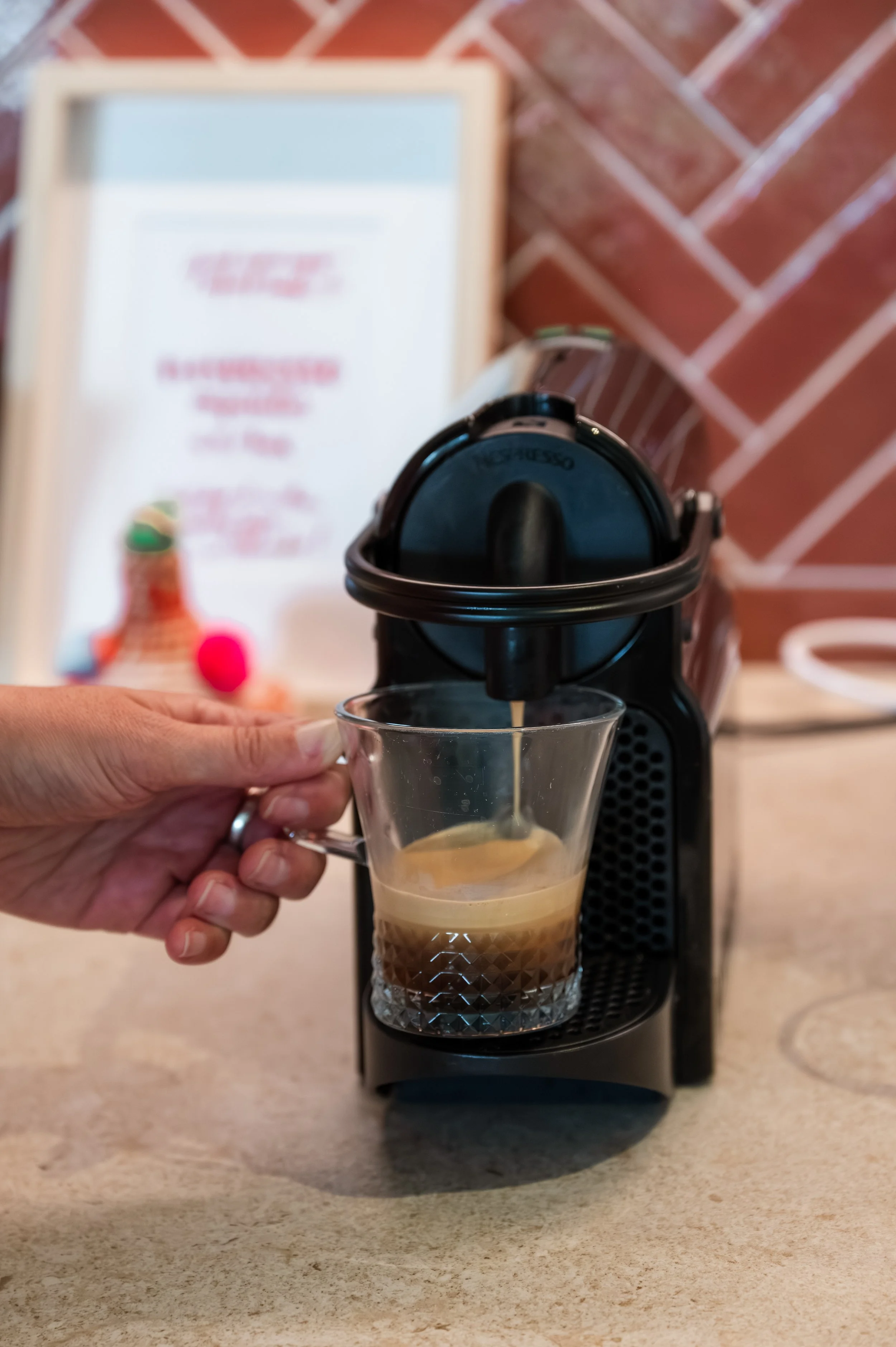 A hand holding a glass of iced coffee being filled by a coffee machine, with a brick wall and a sign in the background.