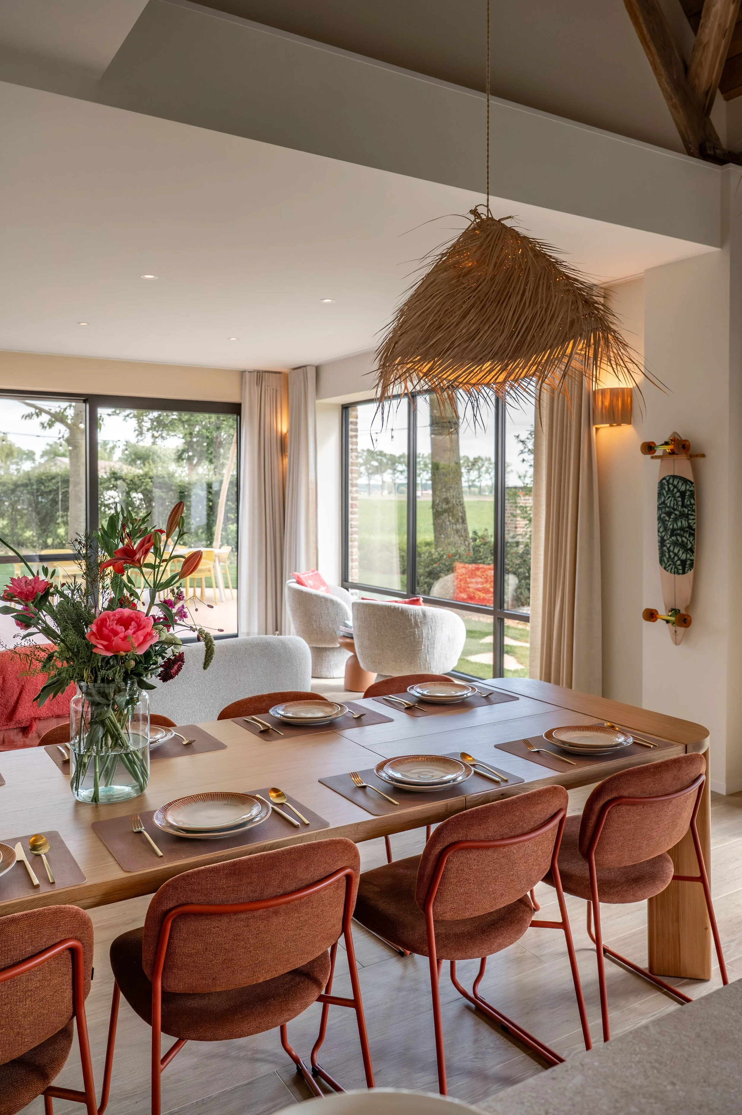 A dining room with a wooden table set for six, with plates, cutlery, and brown placemats. A large vase with pink, red, and purple flowers is at the center of the table. Six upholstered chairs with rust-colored fabric surround the table. Large windows