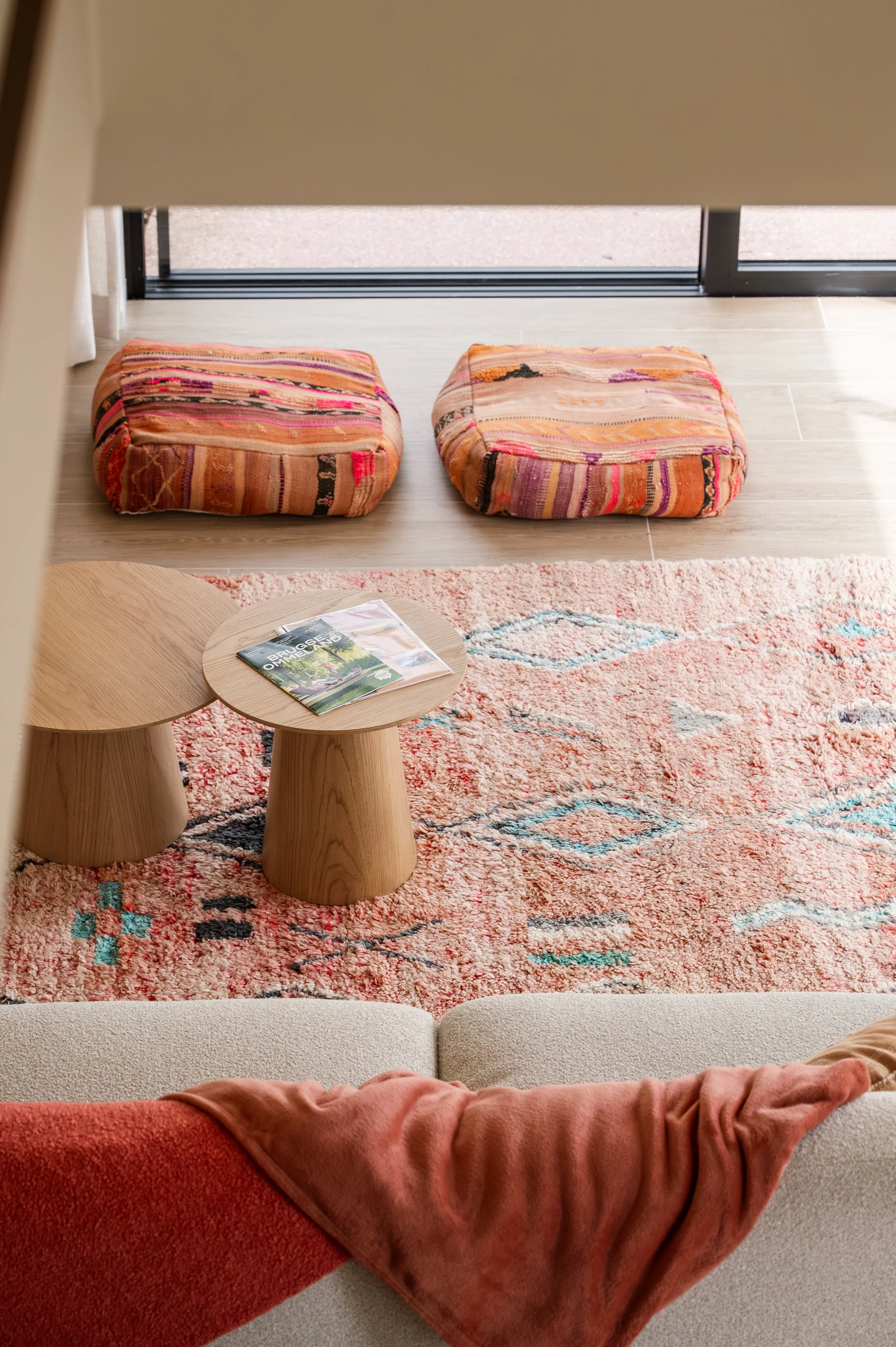 Interior view of a living room with a red plush sofa with a plush pink blanket, a patterned area rug, a wooden coffee table with magazines, and two colorful floor cushions near a sliding glass door leading outside.