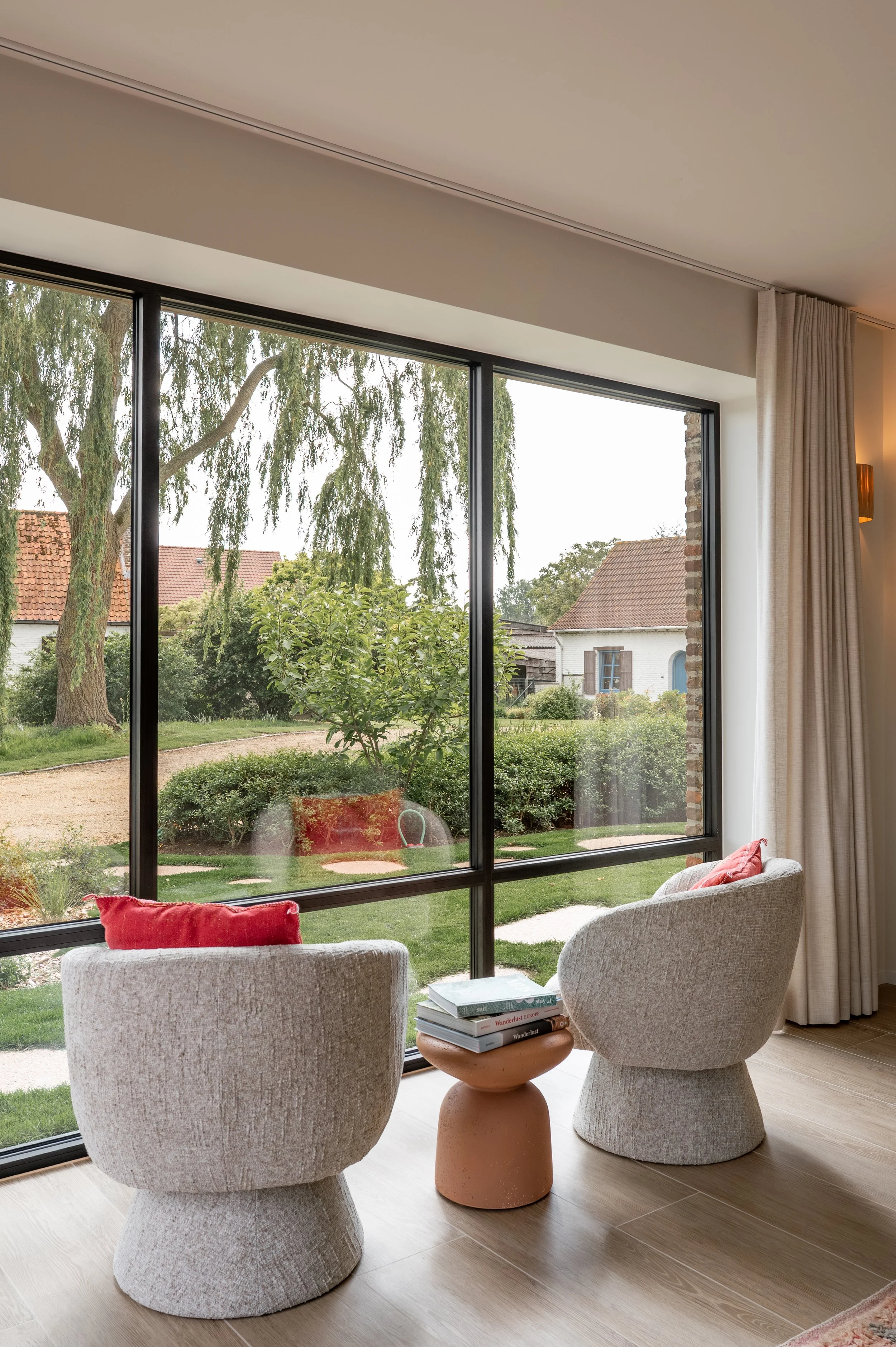Indoor seating area with two beige upholstered chairs and a small round table with books, overlooking a garden with grass, bushes, a large tree, and neighboring houses through large glass windows with white curtains.