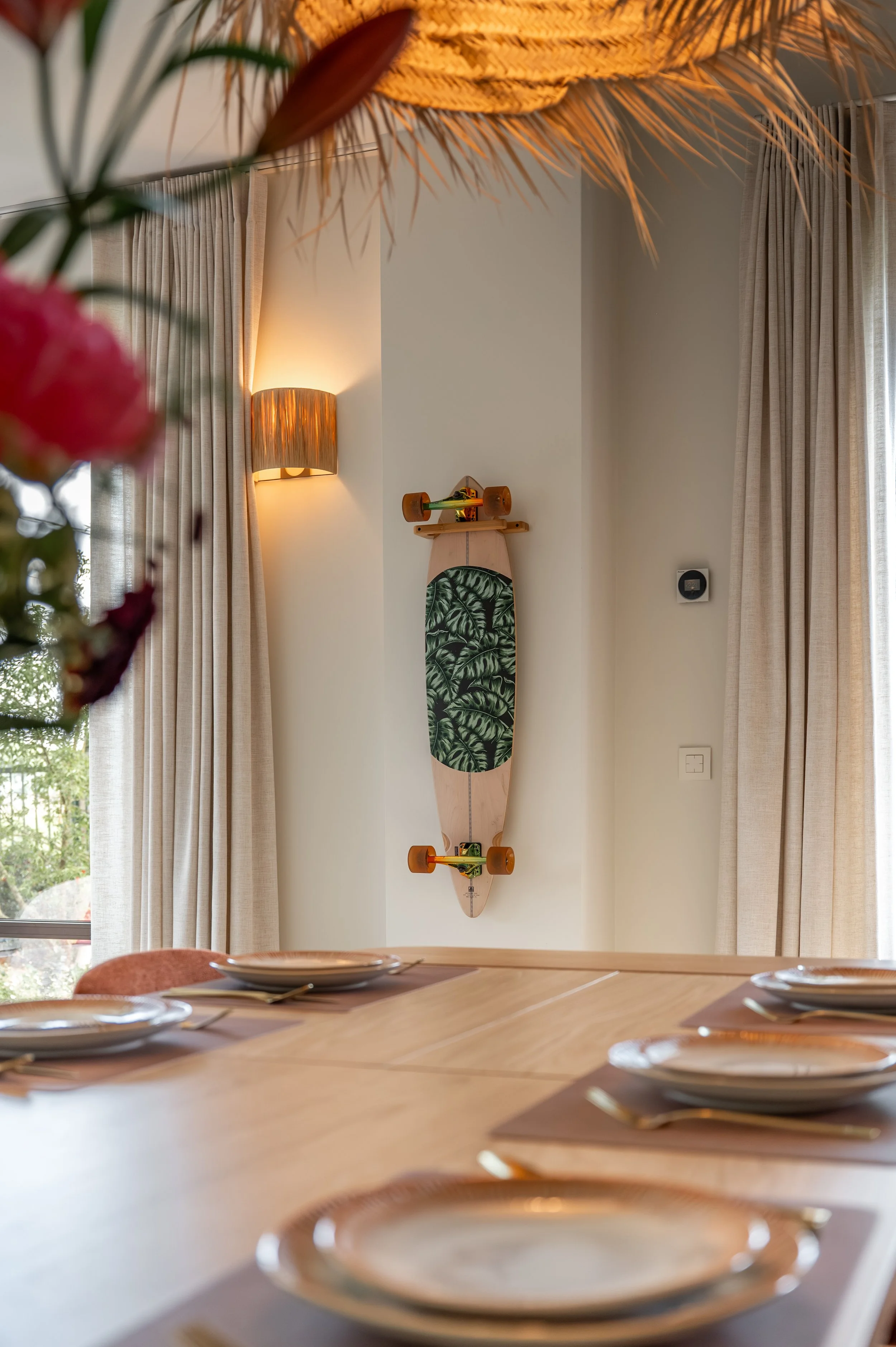 A skateboard mounted on a wall in a room with beige curtains. The skateboard has a tropical leaf pattern on its deck, orange wheels, and visible trucks.