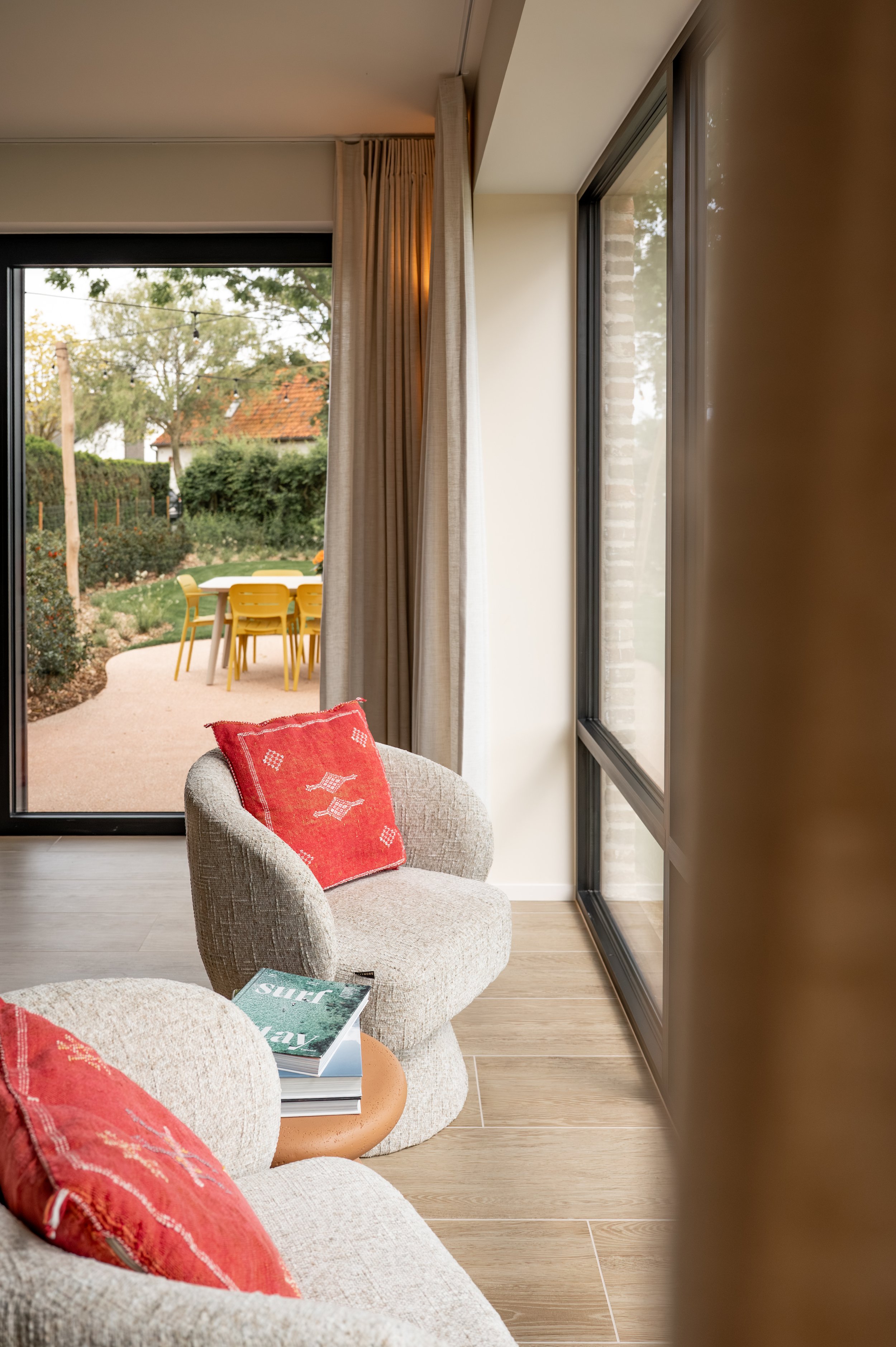 Living room with beige chairs, red pillows, a small round table with magazines, large windows, and sliding glass door leading to an outdoor patio with yellow chairs and a table. There are trees and a house with a red roof outside.
