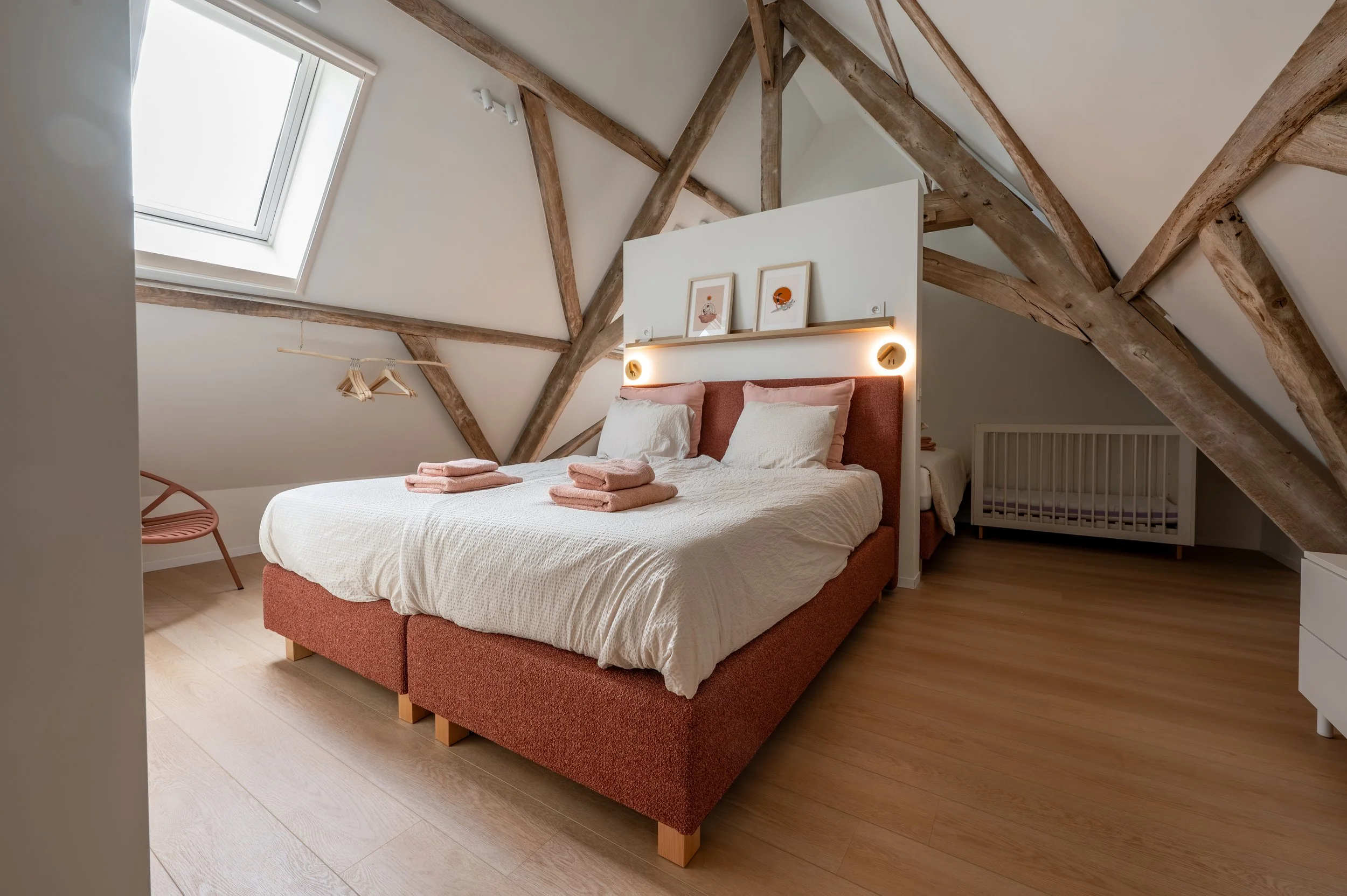 Bedroom with sloped ceiling, wooden beams, skylight window, a bed with pink headboard, white bedding, and folded pink towels, a pink chair, and framed art on the white wall.