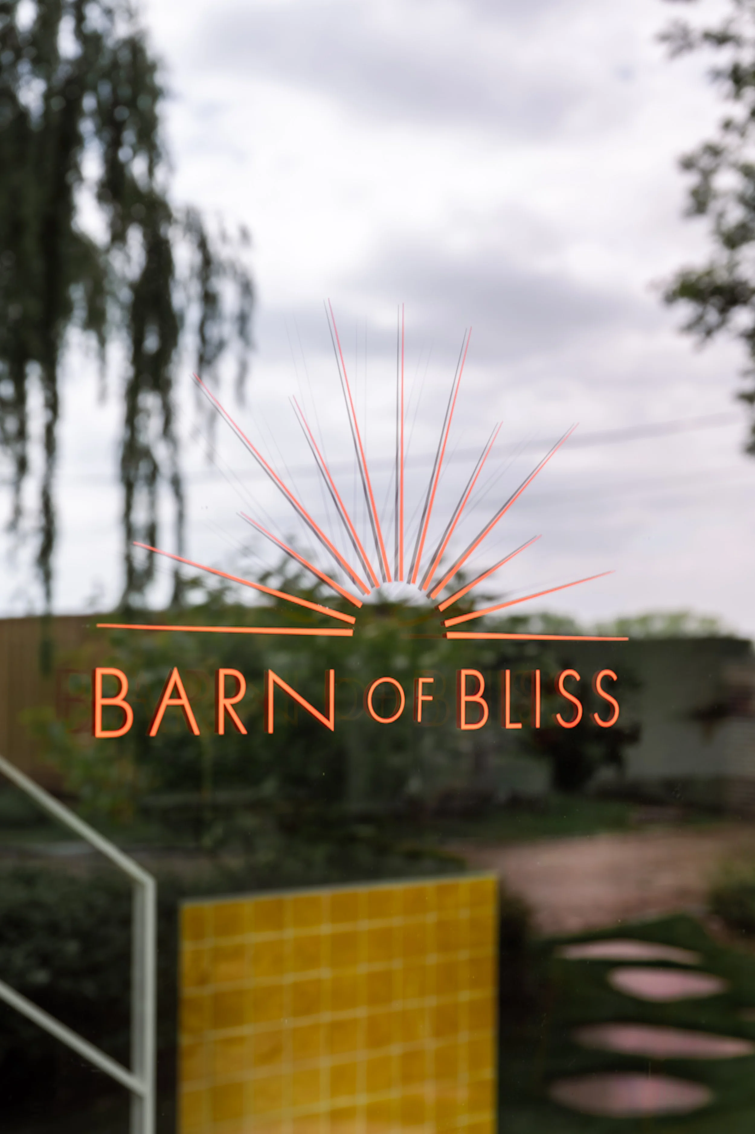 Sign on glass door with the text 'Barn of Bliss' and a sunburst design, reflecting trees and cloudy sky outside.