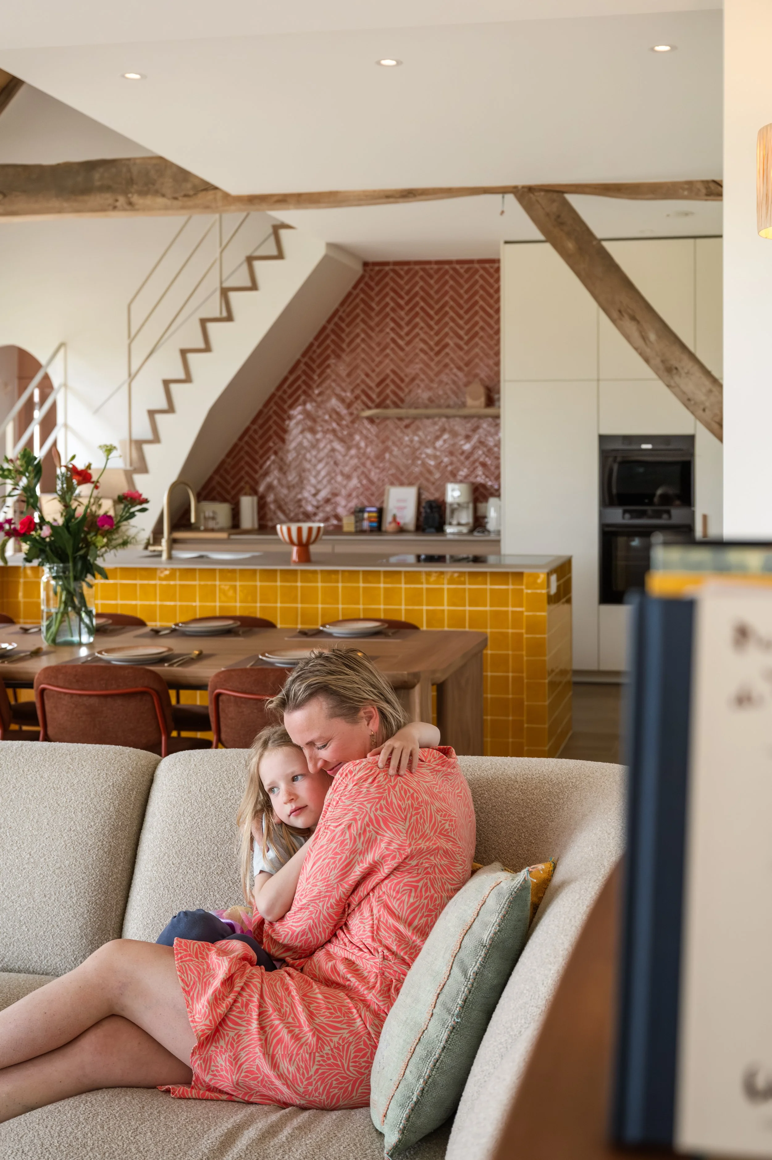 A woman in a patterned pink dress is cuddling a young girl on a beige couch in a cozy, modern kitchen with exposed wooden beams, red brick backsplash, and yellow tile accents.