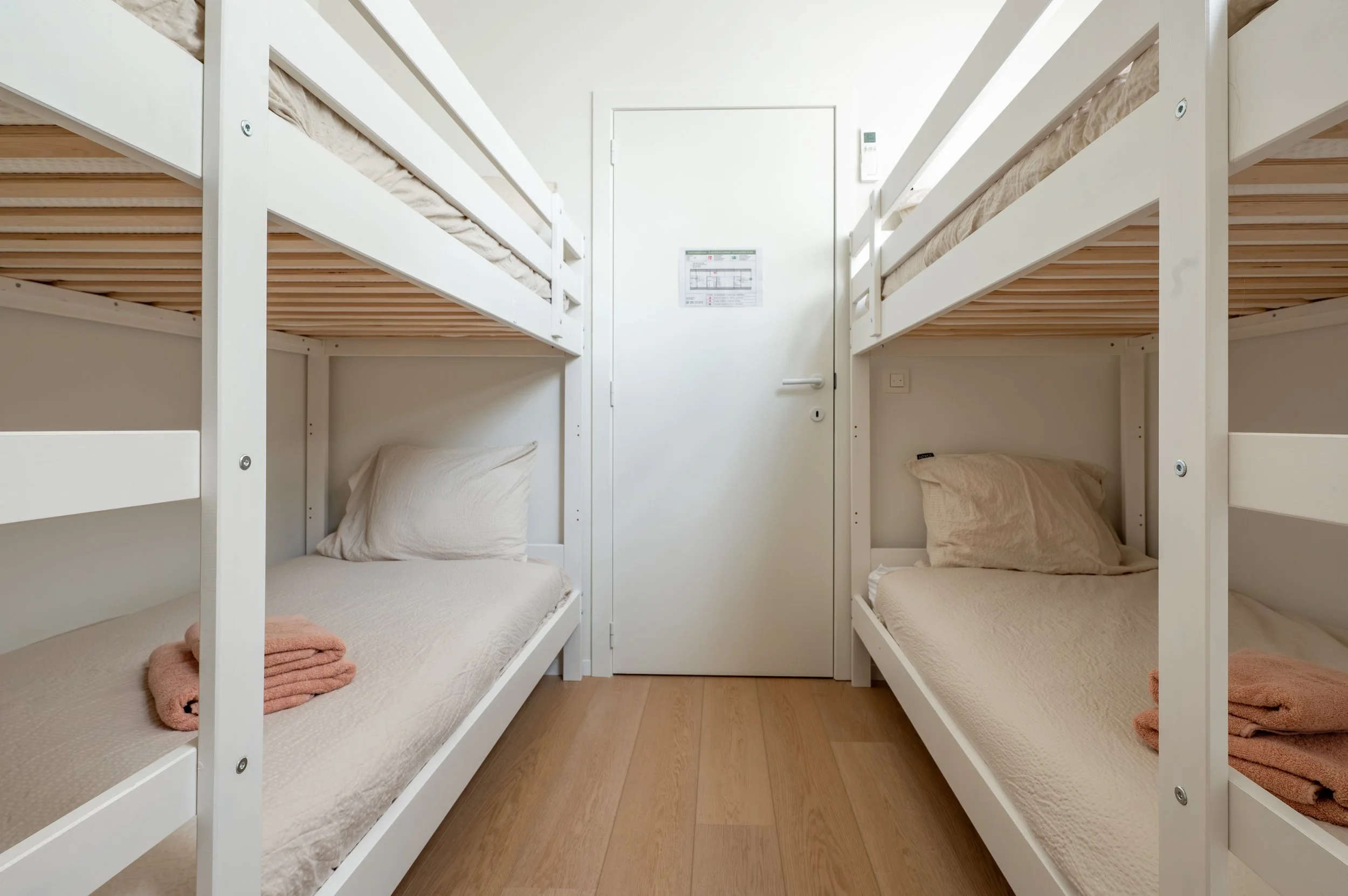A small room with two sets of white bunk beds against the walls, each with beige pillows and bedsheets. Towels are folded at the foot of each bed, and a closed white door is in the center of the wall.