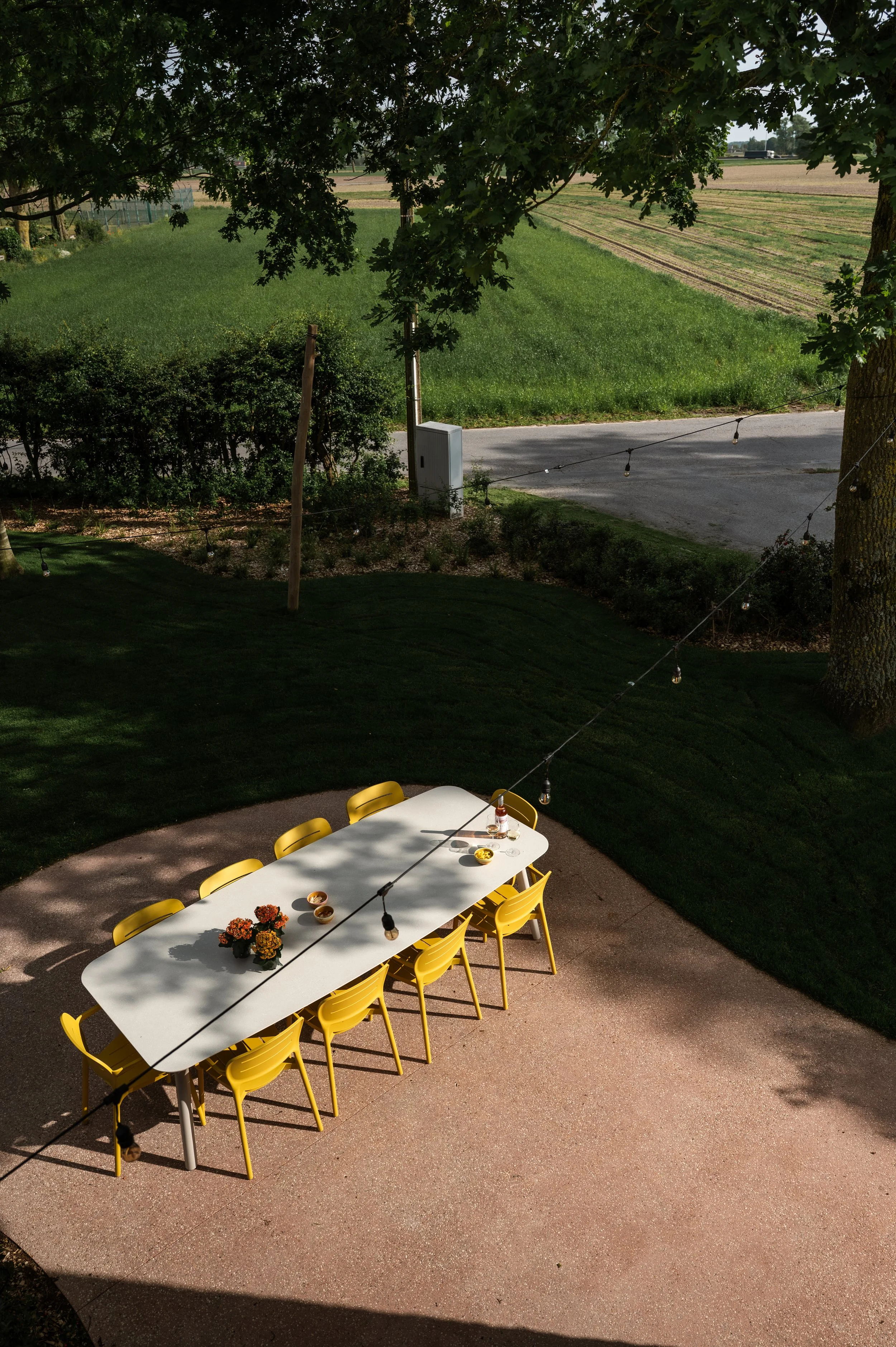 A white outdoor table with yellow chairs, decorated with potted flowers, set on a patio with a backyard and green lawn, with trees and fields in the background.