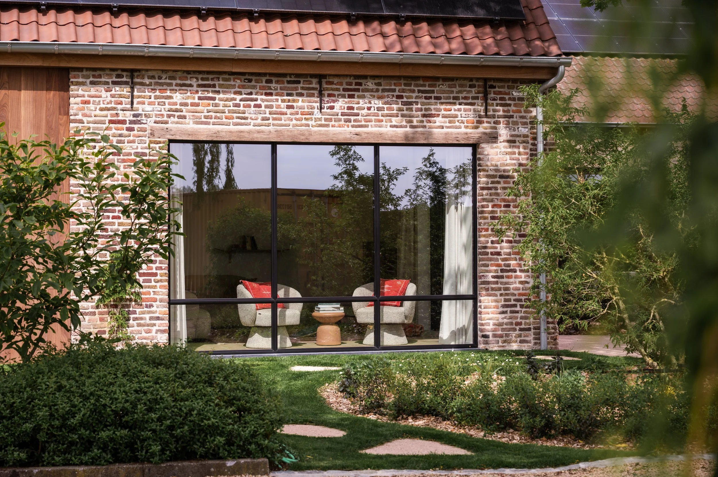 A house with a brick exterior, large glass window, and a red tiled roof with solar panels. Inside, two beige armchairs with red pillows and a small round wooden table are visible through the window. There are lush green bushes and lawn outside.