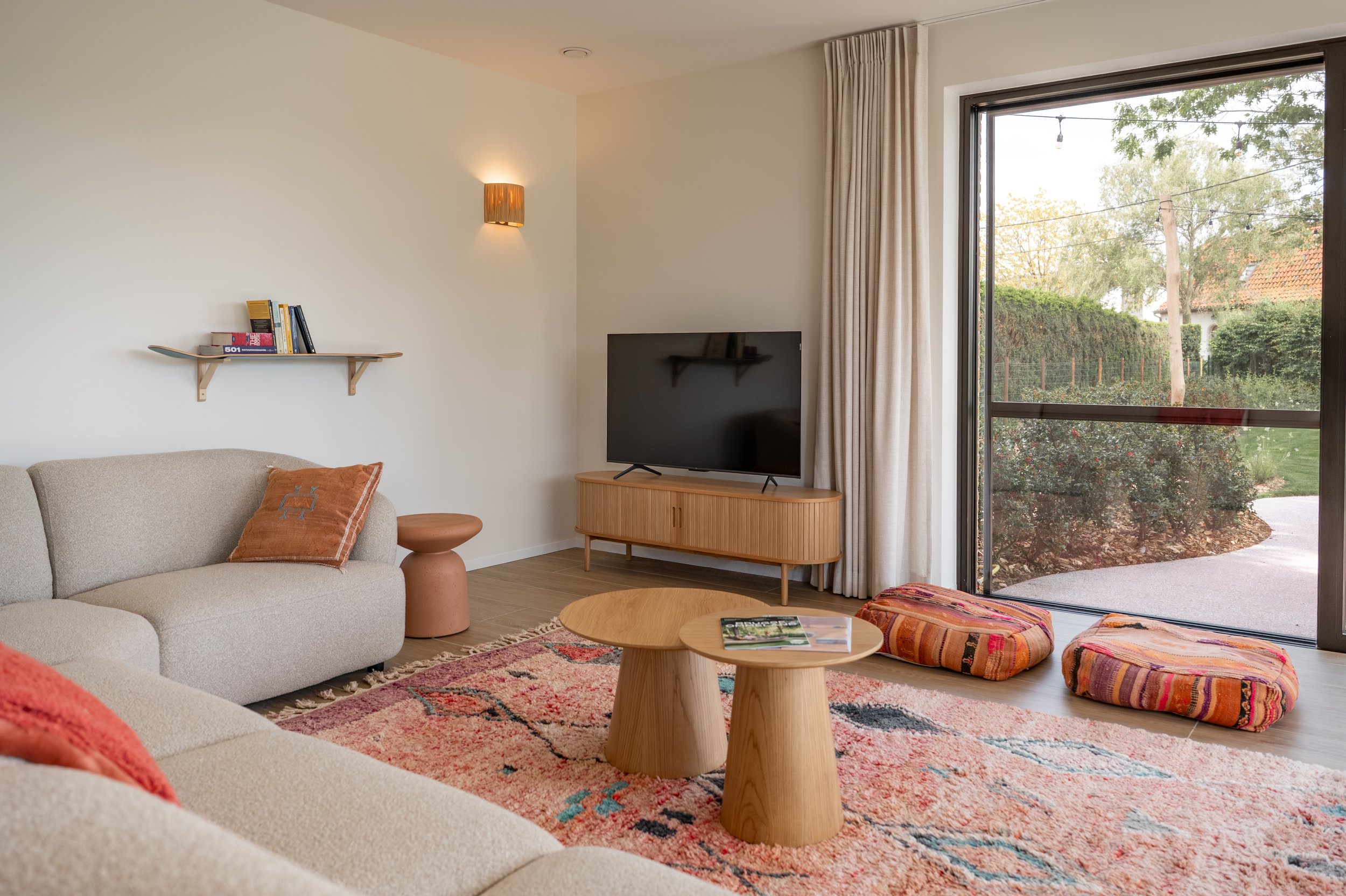 Living room with beige sofa, orange pillows, round wooden coffee table, colorful rug, black flat-screen TV on wooden stand, cream curtains, floor-to-ceiling window showing trees outside, wall sconce, and books on a wall shelf.