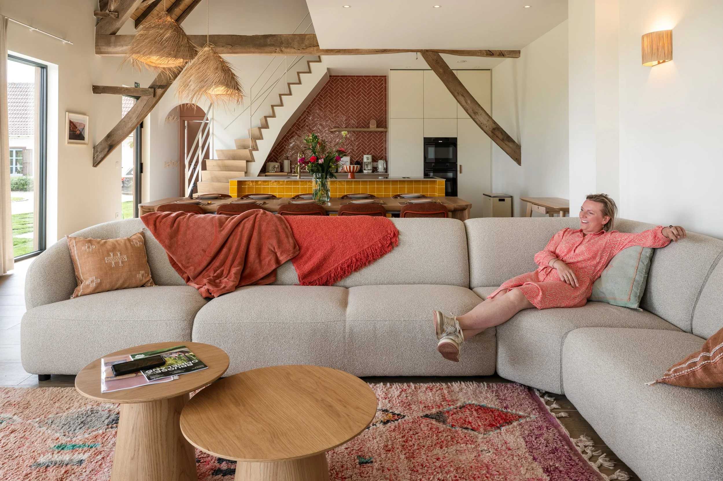 A woman relaxing on a light-colored sofa in a modern living room with a rustic design, pink floral dress, and blonde hair, smiling. In front of her is a round wooden coffee table with magazines. Behind her is a kitchen area with a red brick backsplas