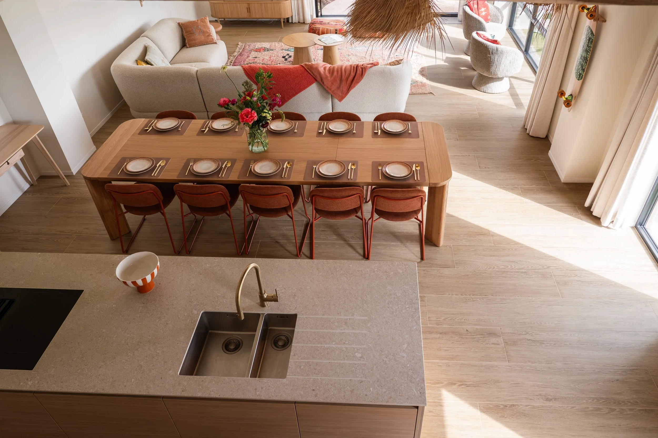 A modern open-plan living and dining area with a wooden dining table set for ten, featuring a flower arrangement, and a light-colored sofa with throw pillows. The space has wood flooring, large glass windows with curtains, and contemporary decor incl