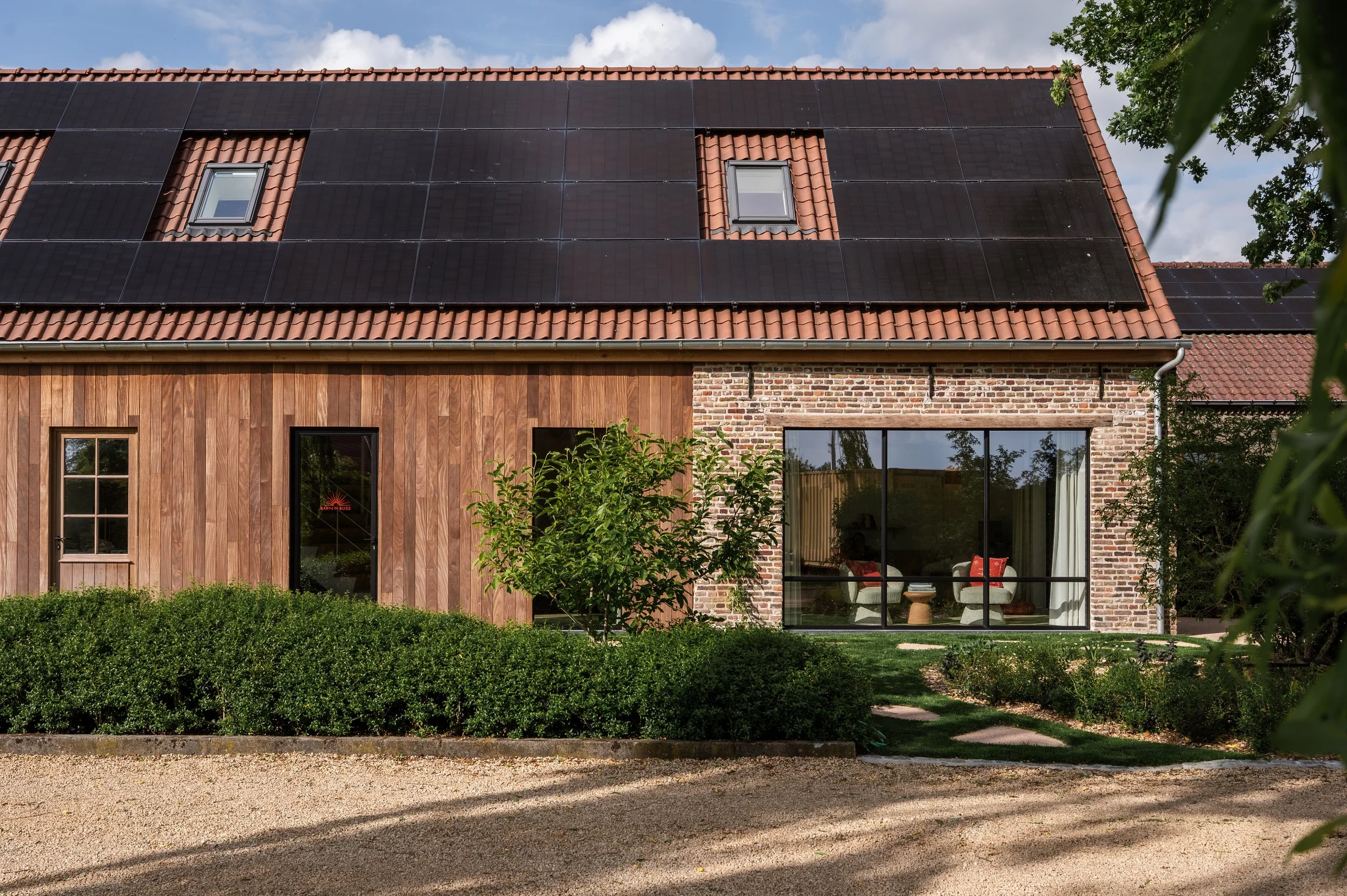A house with a brick lower wall and a wooden upper wall, large glass sliding doors, and a roof covered with black solar panels. There are two small dormer windows on the roof, and greenery, including bushes and a tree, surrounds the house. The sky is