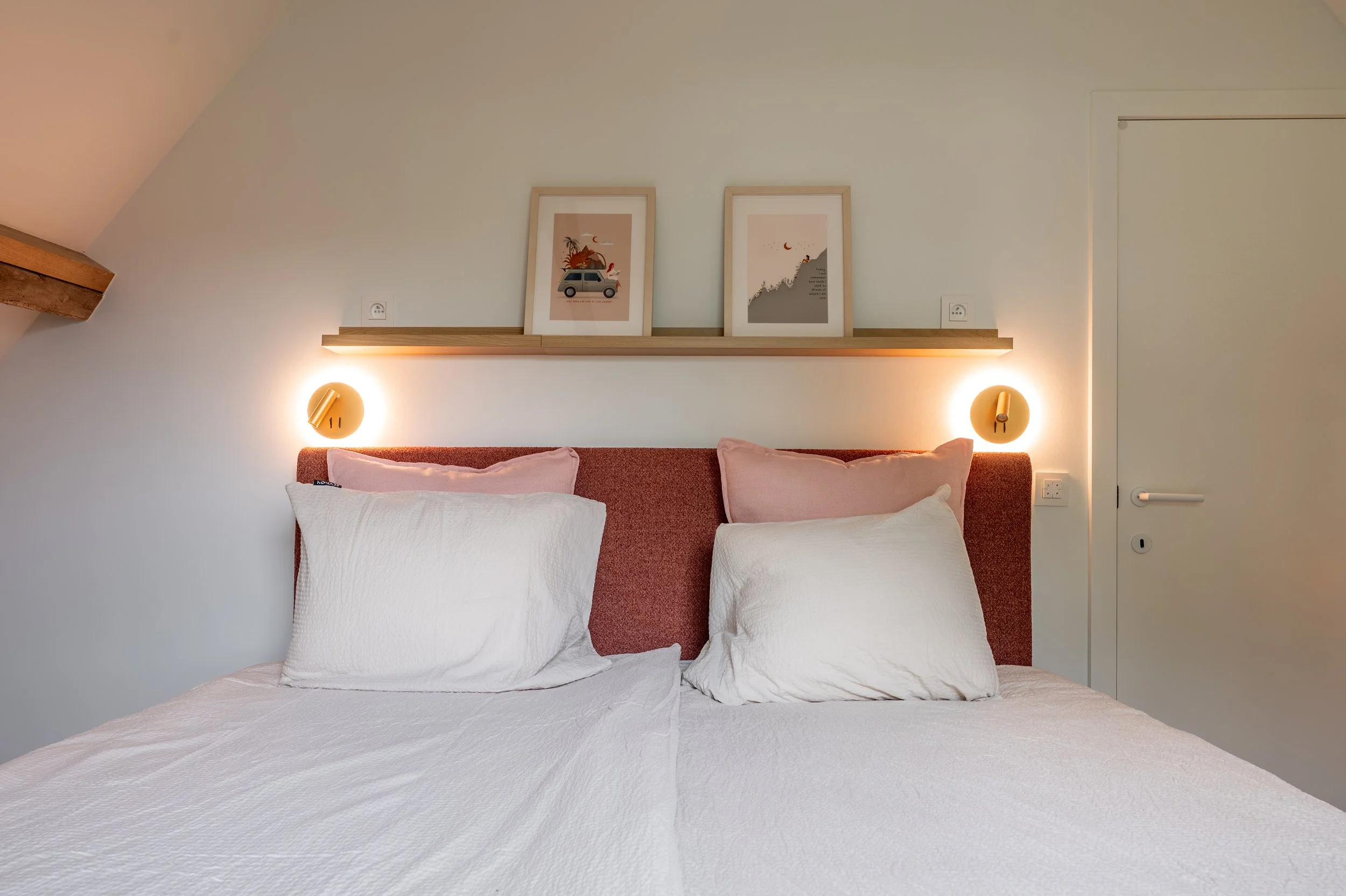 A neatly made bed with white bedding and two pillows, a brick-colored headboard, two circular wall-mounted lights on either side, a wooden shelf with framed artwork above the bed, and a white door to the right.