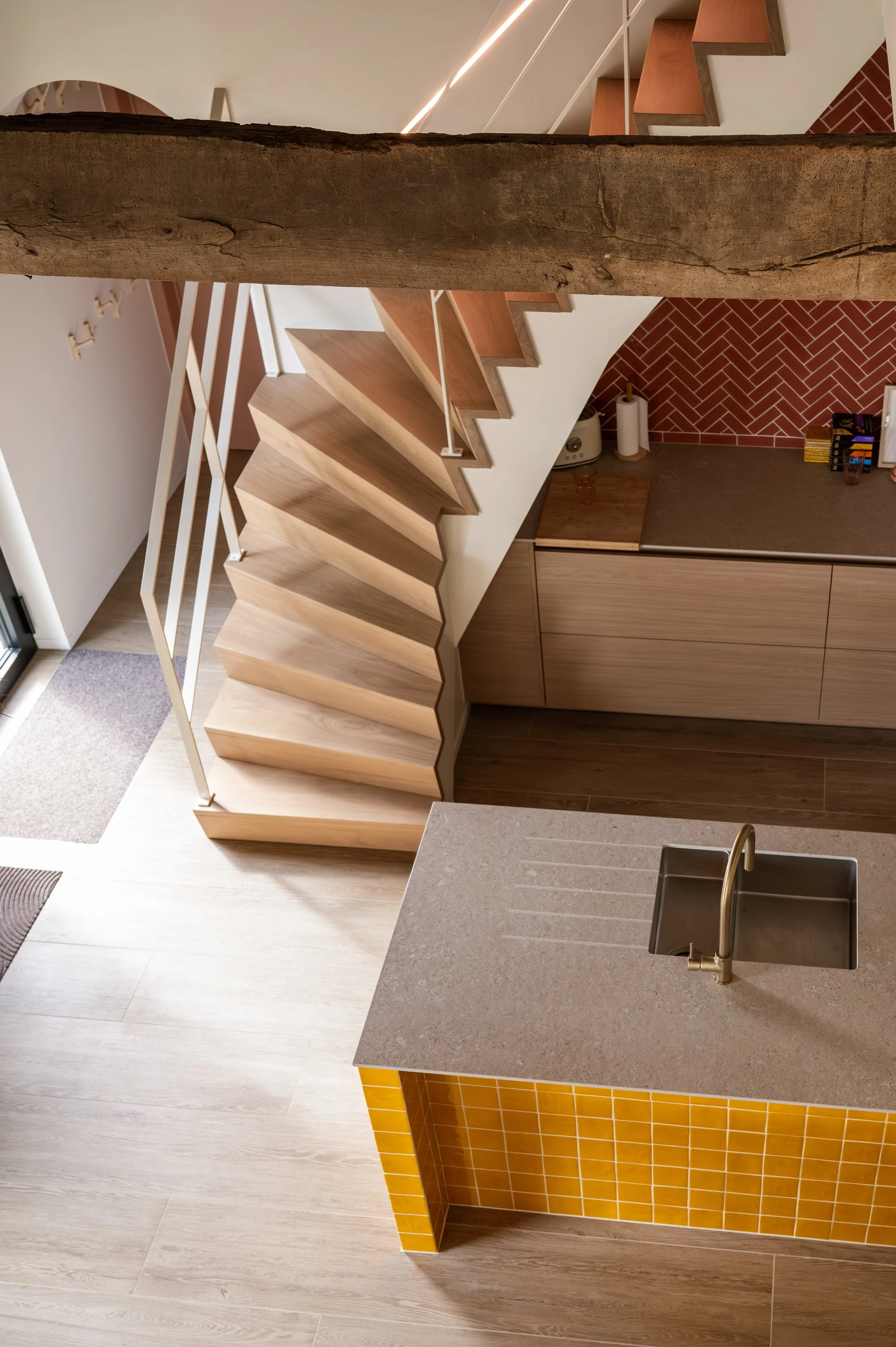 Interior view of a modern kitchen with wooden stairs and a support beam. The kitchen features a beige countertop with a brass faucet, yellow tile backsplash, and dark brown cabinets. There is a red brick wall, paper towel roll, and various small item