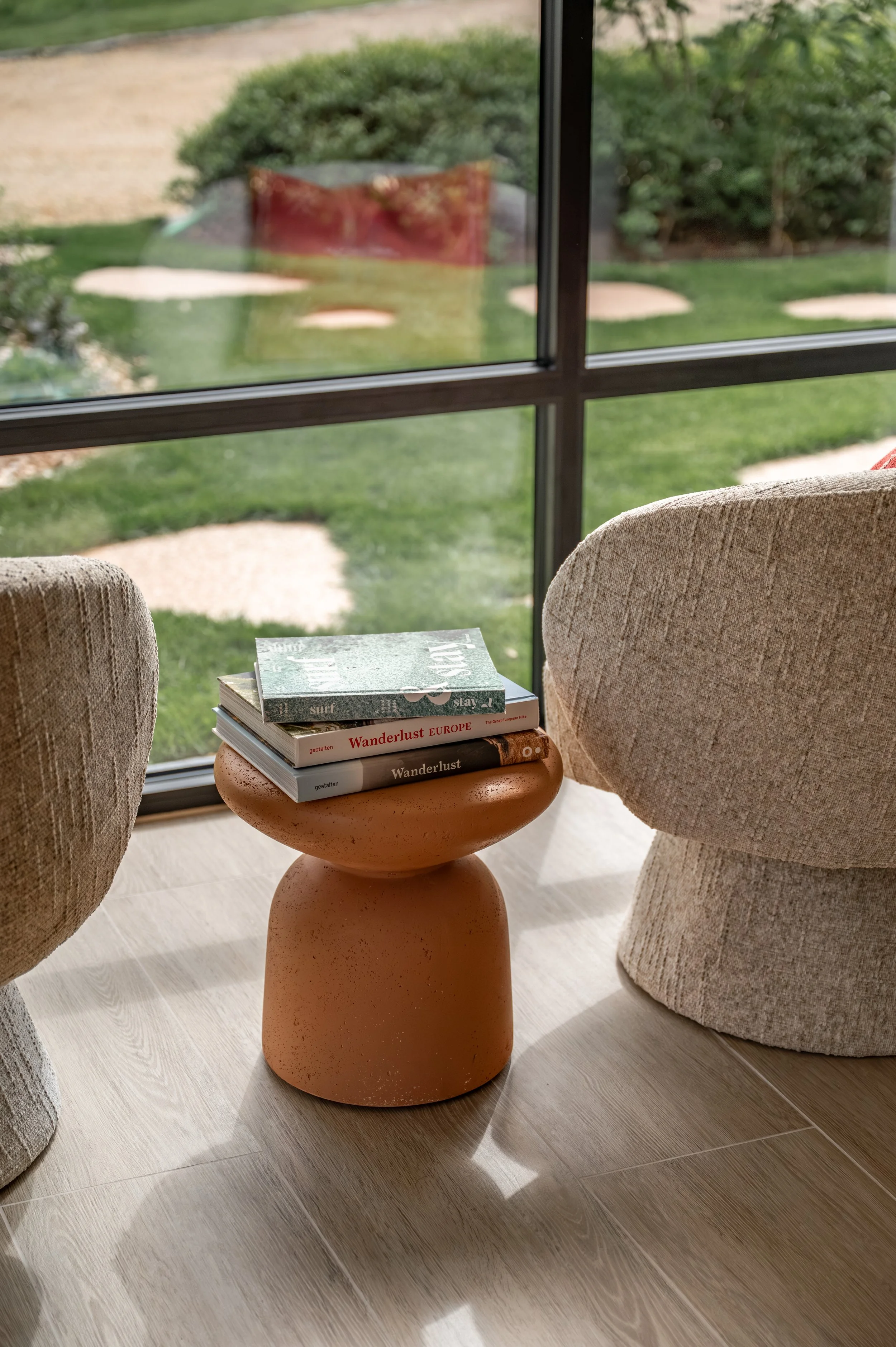 A small round clay side table with a stack of books on top, two upholstered armchairs on either side, and a large window showing a garden with bushes and stepping stones outside.
