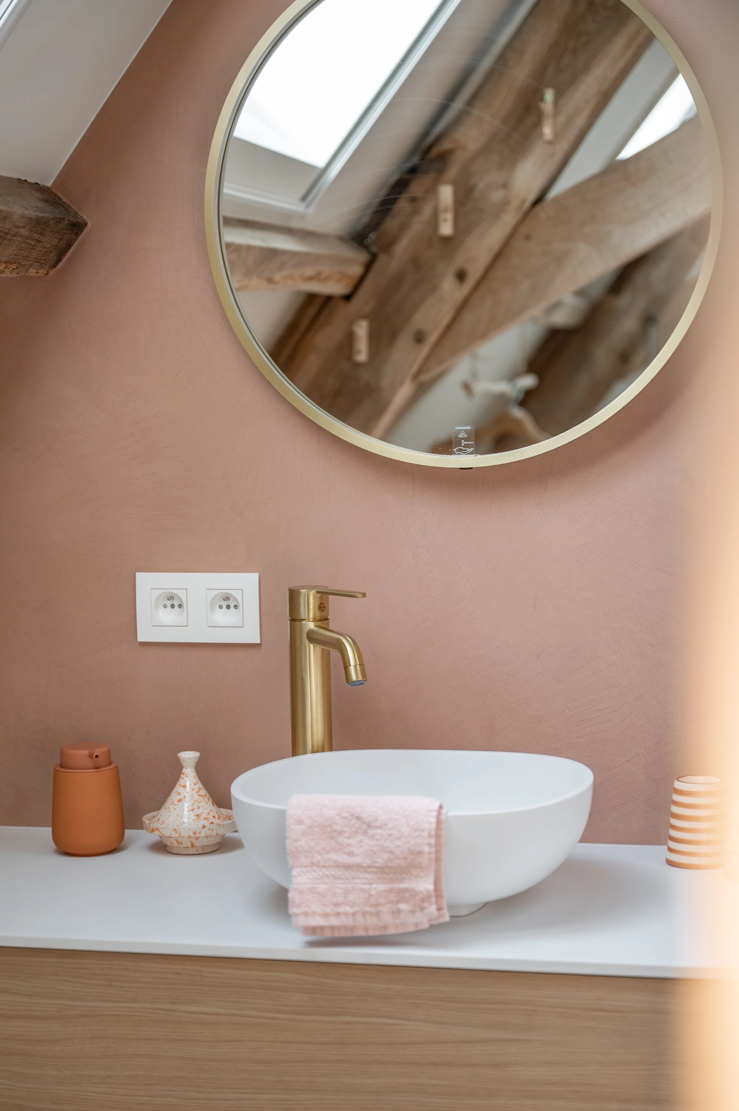 Modern bathroom with a round white vessel sink, gold faucet, pink walls, and decorative vases in terracotta, patterned ceramic, and striped designs. A mirror is reflected in the picture.