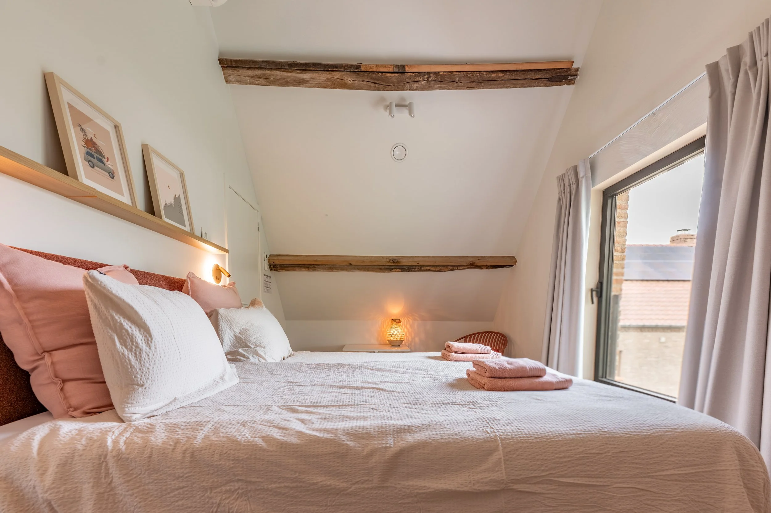 A cozy bedroom with a bed dressed in pink and white bedding, framed artwork on a wooden ledge, and exposed wooden beams on the ceiling. Large window with white curtains lets in natural light, and a small lamp glows in the corner.