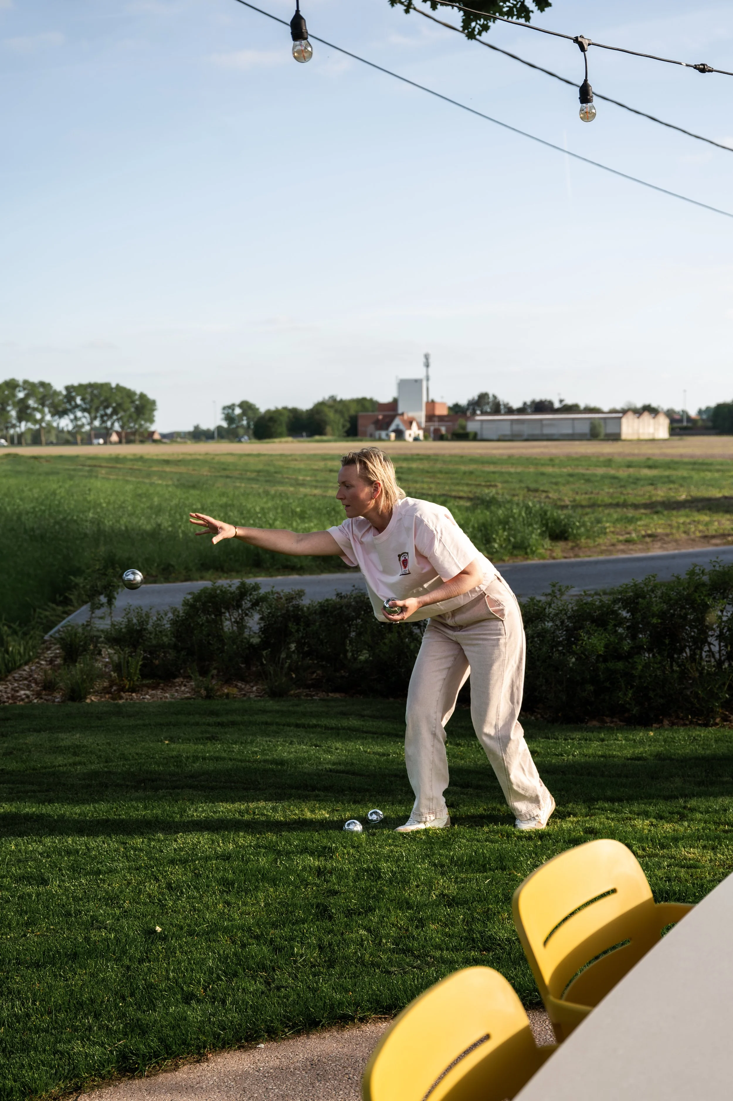 A woman is playing outdoor lawn game with metallic balls on a grassy area in the late afternoon or early evening, with a school or community building and trees in the background, and string lights overhead.