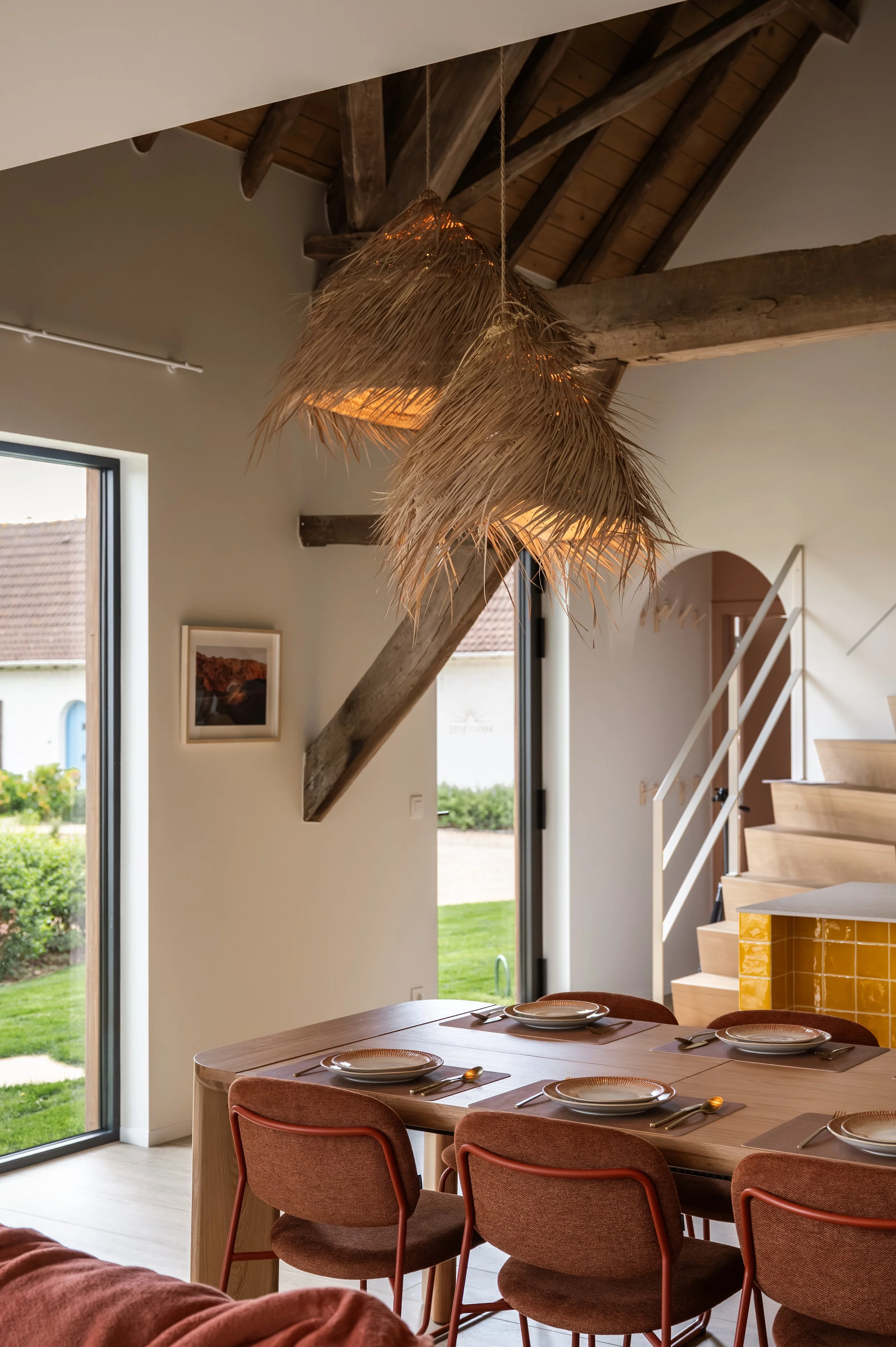 A dining area with a wooden table set with plates and cutlery, surrounded by upholstered chairs, next to large glass doors showing a view of a green lawn outside. Hanging from the ceiling are three woven, straw pendant lights, and part of a staircase