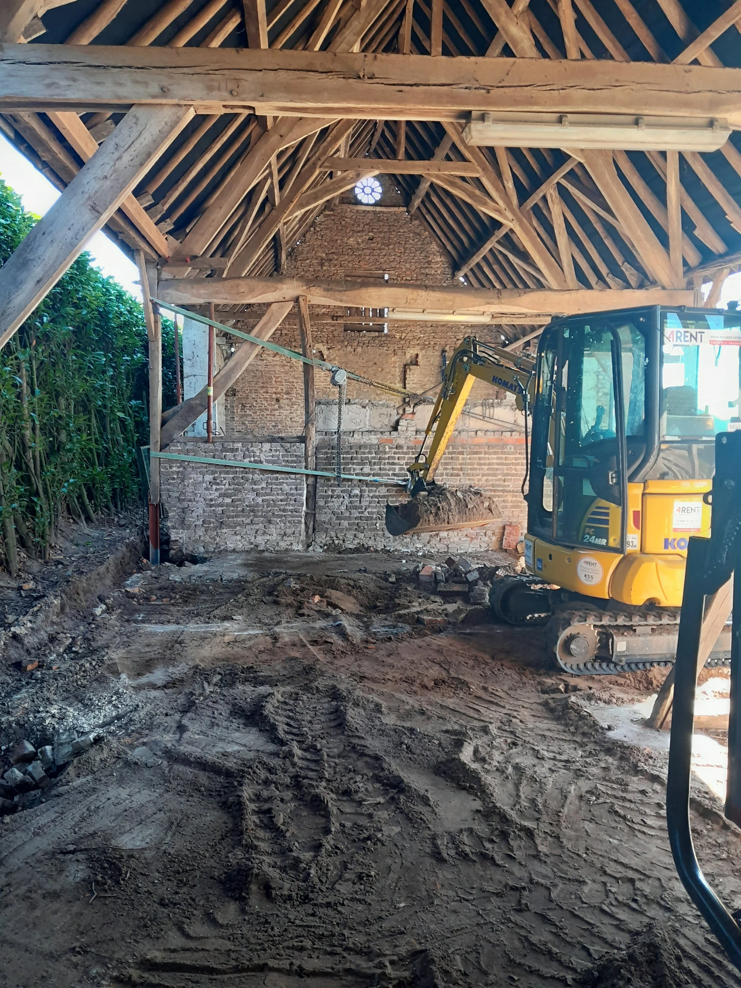 Construction site inside a partially built structure with a small excavator and a dirt floor, unfinished brick walls, and wooden roof framework.