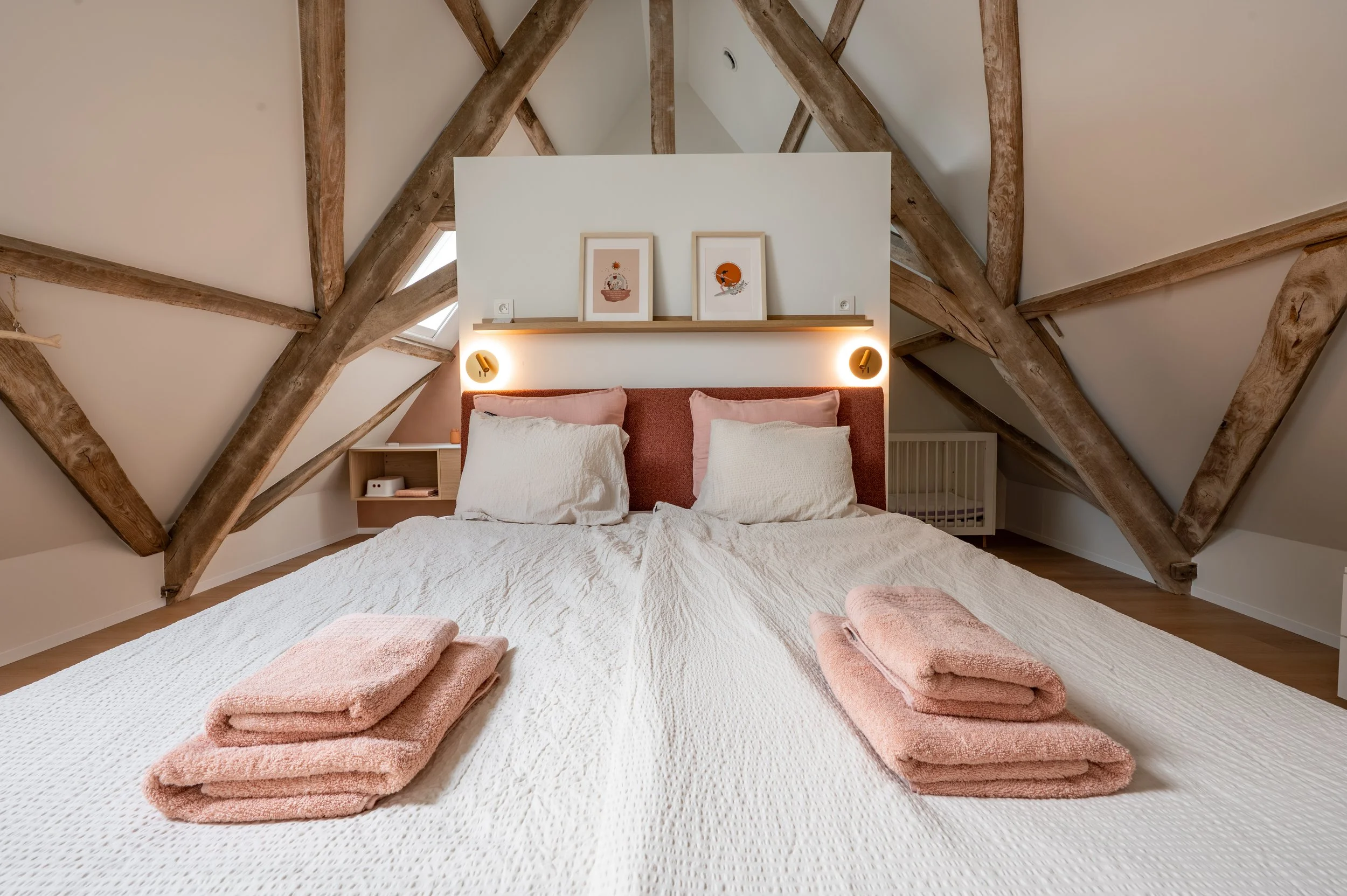 A neatly made bed with a beige textured blanket, pink pillows, and folded pink towels at the foot. Above the bed, a white wall features two framed art prints and built-in lighting. The room has exposed wooden beams on the sloped ceiling.