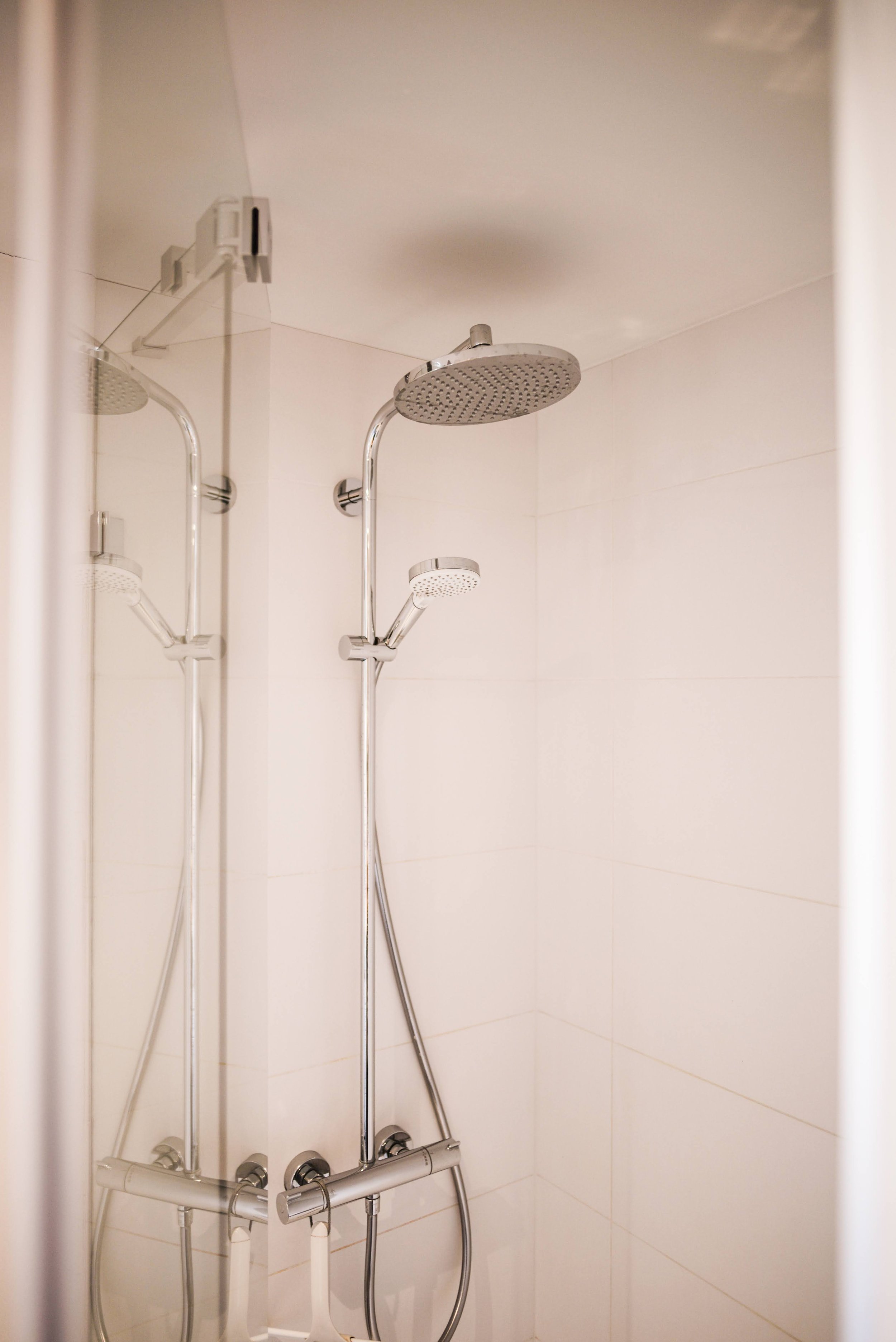 A shower with two shower heads, one large and one smaller, installed with silver fixtures on a white tiled wall.