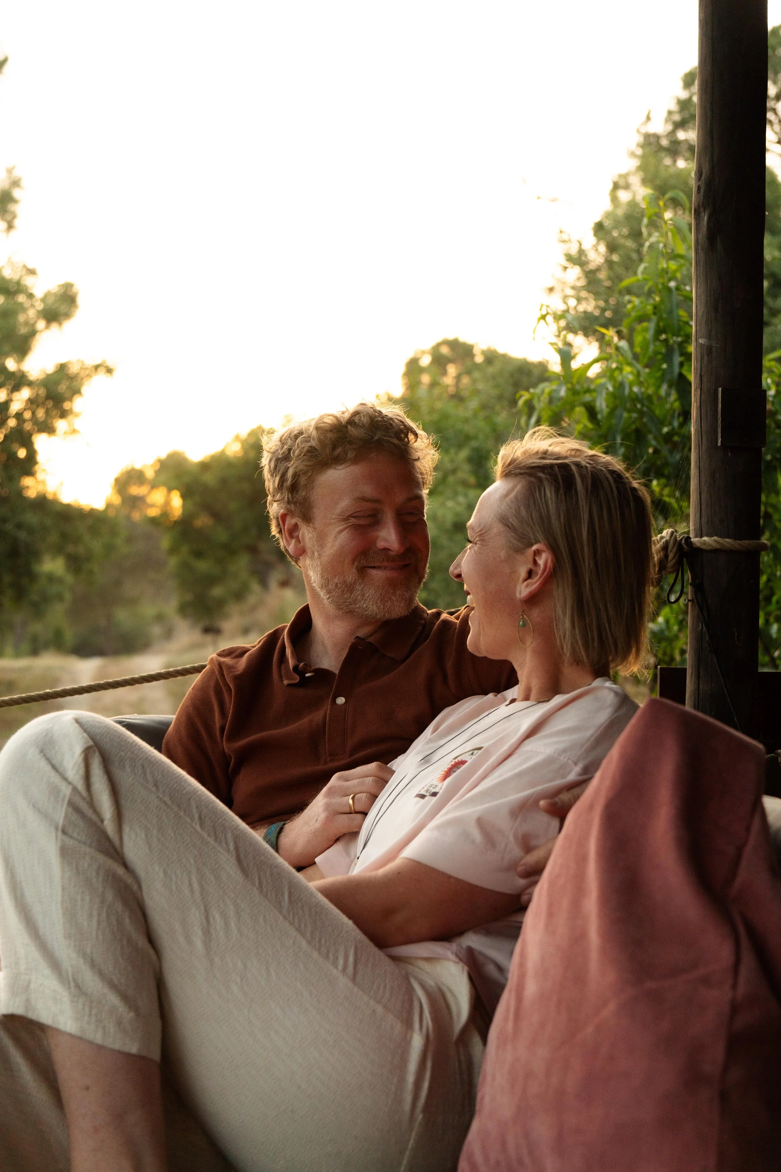 A man and a woman sitting close together outdoors during sunset, smiling and looking at each other.