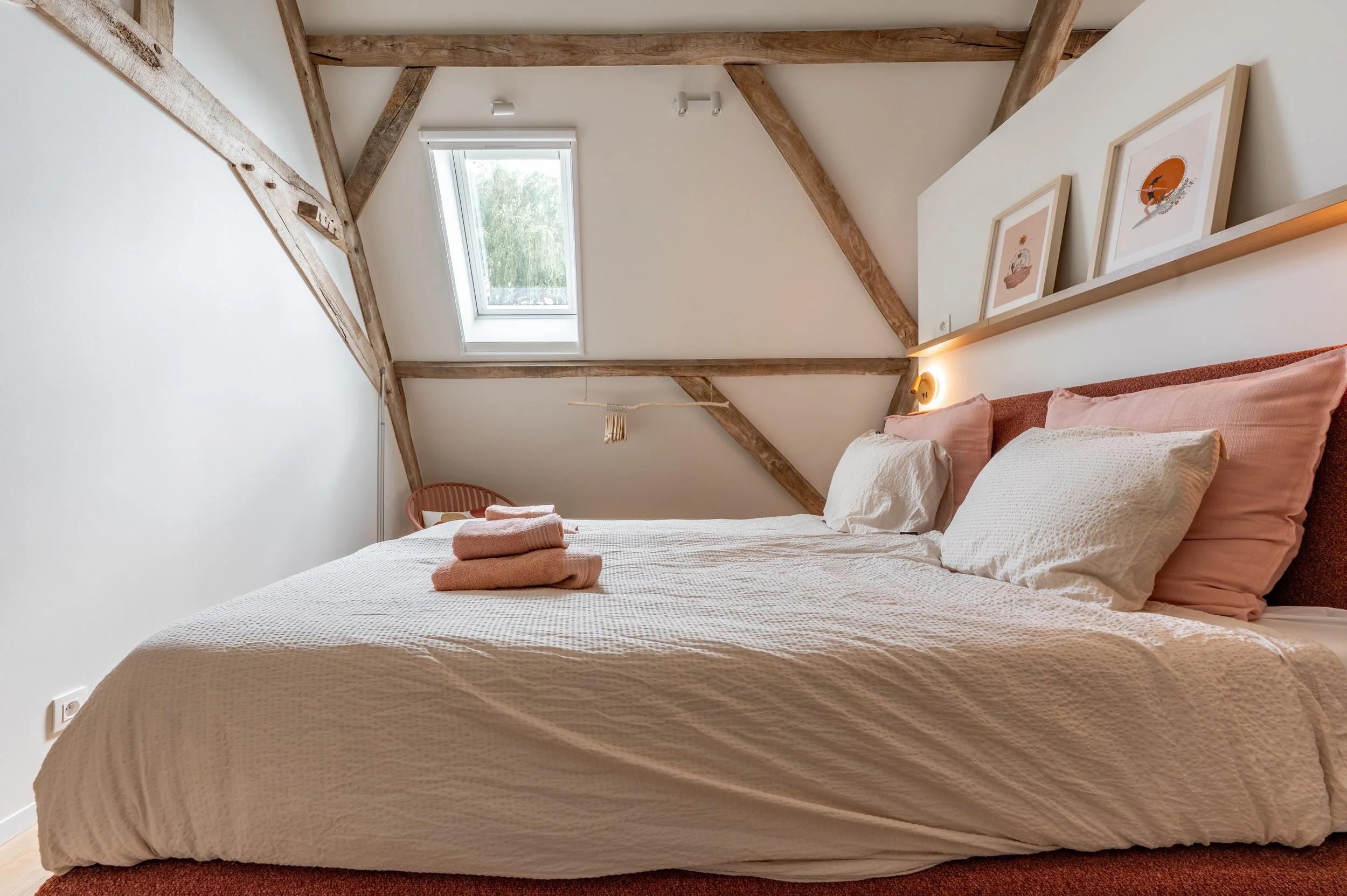 A cozy bedroom with a sloped ceiling, wooden beams, a skylight window, a bed with light-colored bedding, and framed artwork on the wall.