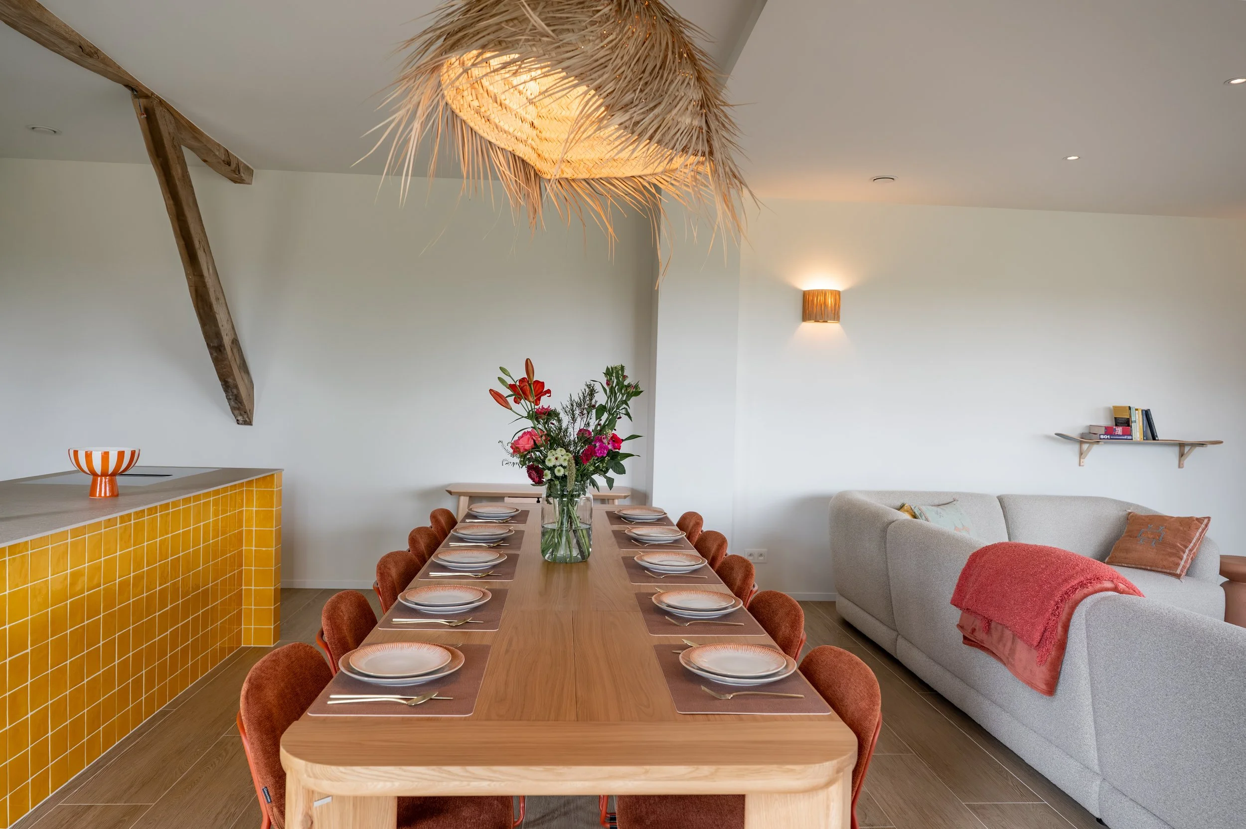 Dining area with a long wooden table set with plates, glasses, and cutlery, surrounded by orange upholstered chairs, a large glass vase with colorful flowers at the center, and a modern hanging light fixture made of woven natural fibers.