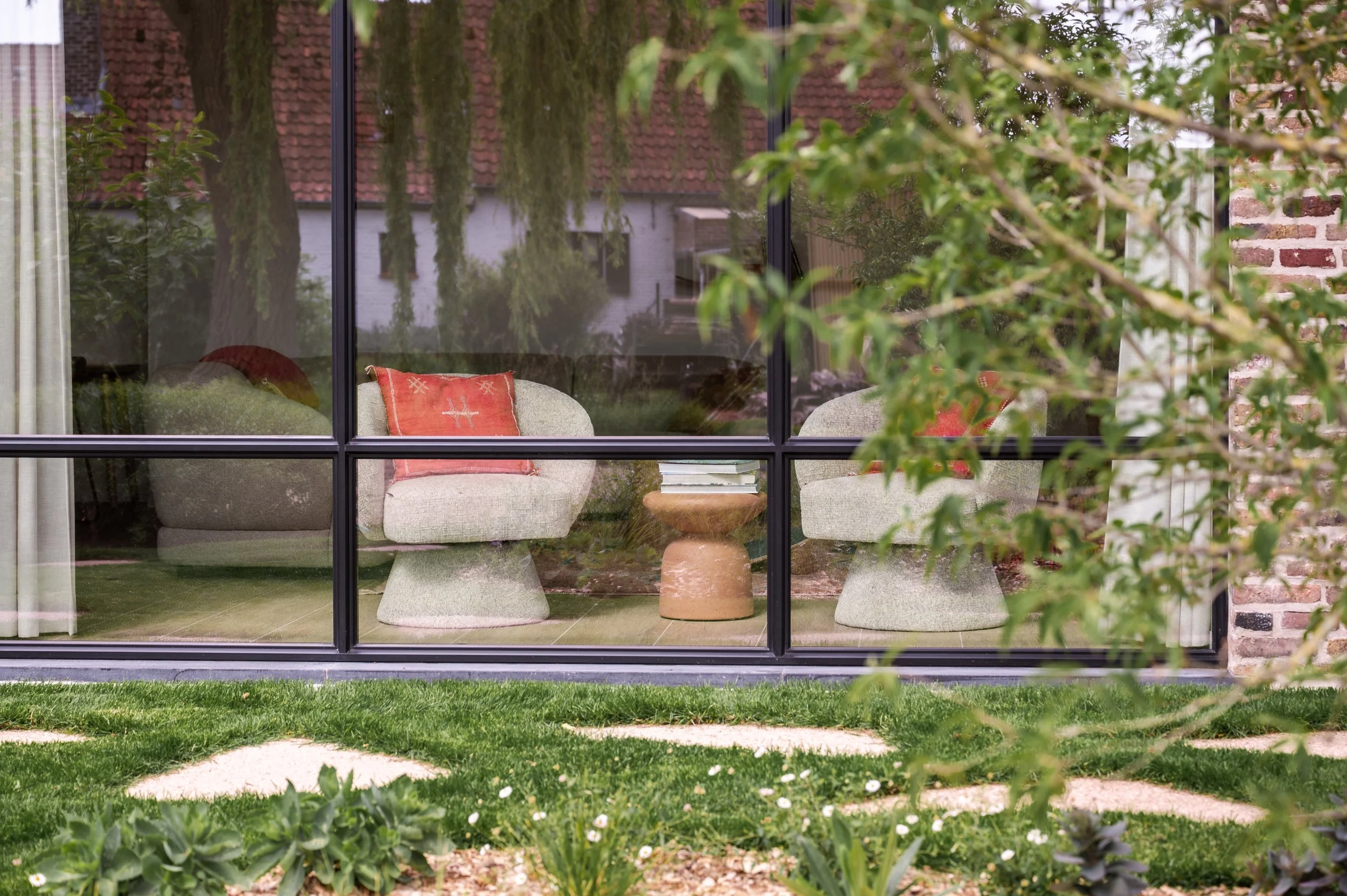 View of a modern house interior with large glass windows showing a sitting area with two chairs, a cushion, and a small table inside, seen through the window from a lush garden outside.